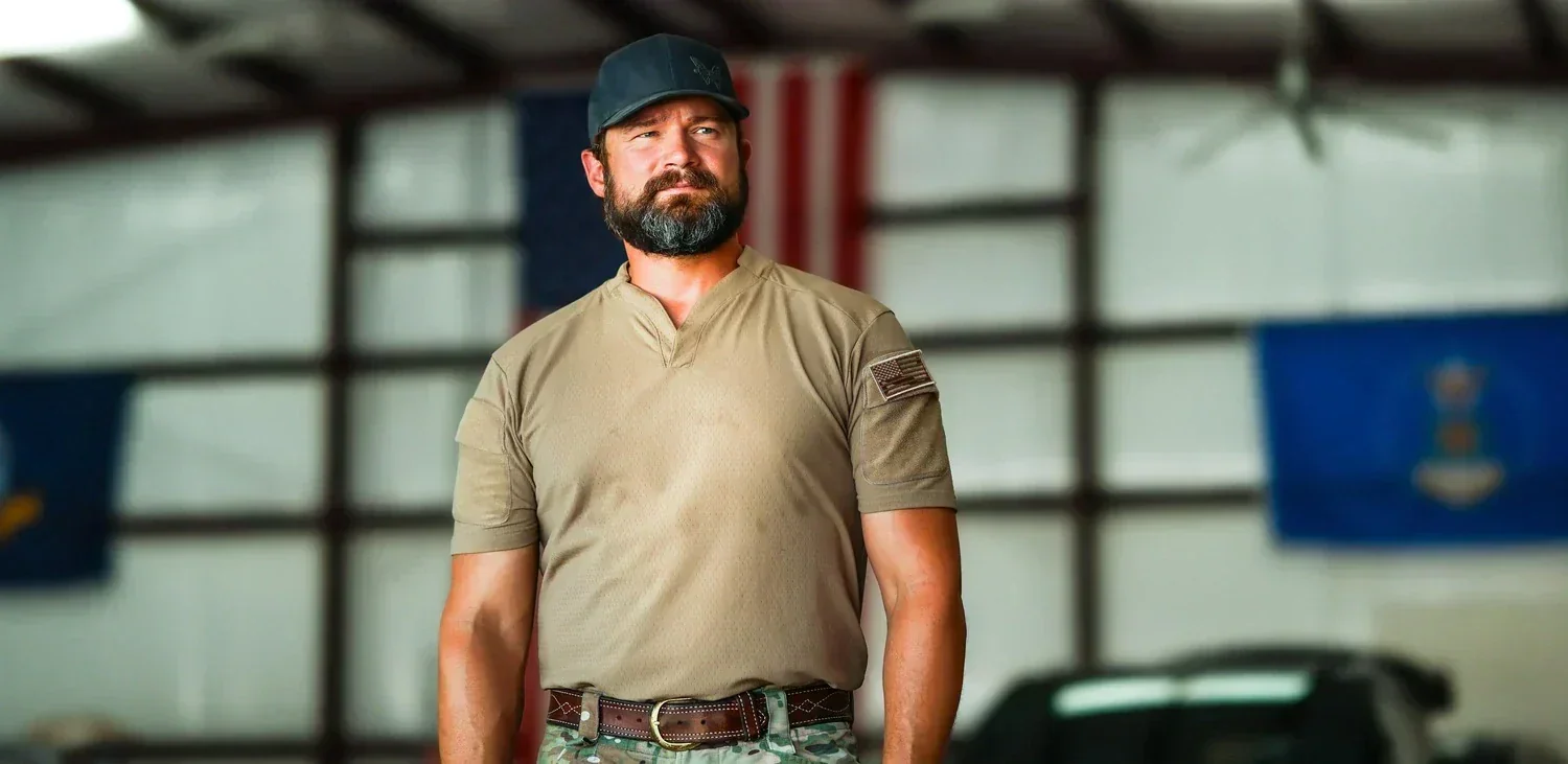 A bearded man wearing a khaki shirt and a baseball cap standing indoors with flags in the background.