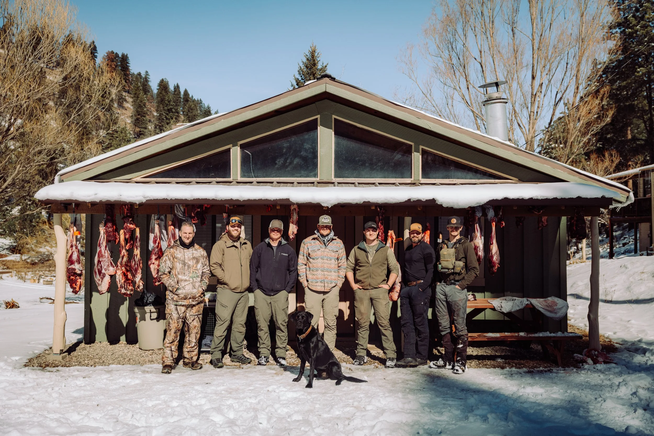 Group of eight men and a black dog standing in front of a shed with hanging meat, in a snowy outdoor setting.