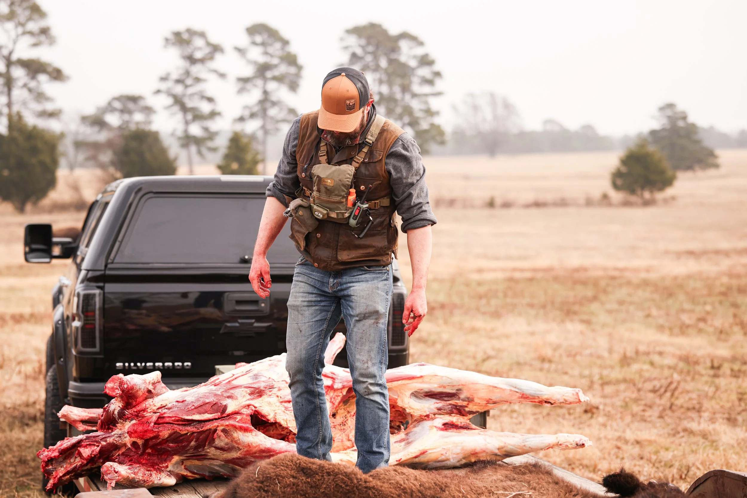 A man dressed in outdoor clothing with a vest and baseball cap stands in an open field next to a large carcass of an animal, likely a deer or similar large game, lying on a trailer. A black Chevrolet Silverado truck is parked behind him, and the background features trees and a cloudy sky.