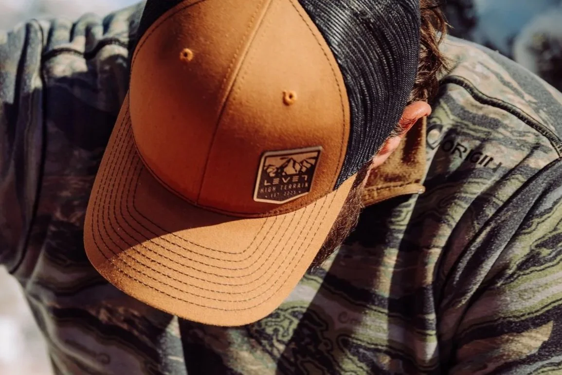 Close-up of a person wearing a camouflage jacket and a two-tone tan and black baseball cap with a mountain logo and text, looking down.