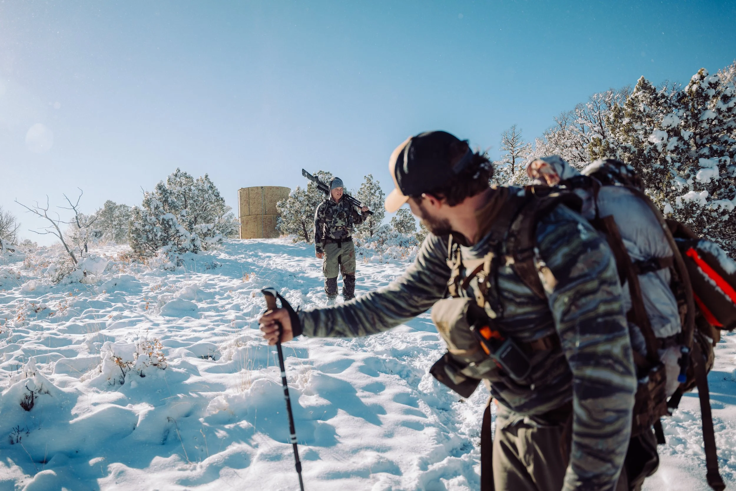 Two hikers traversing a snow-covered landscape with trees and a cylindrical structure in the background, one with a walking pole and the other carrying a rifle.