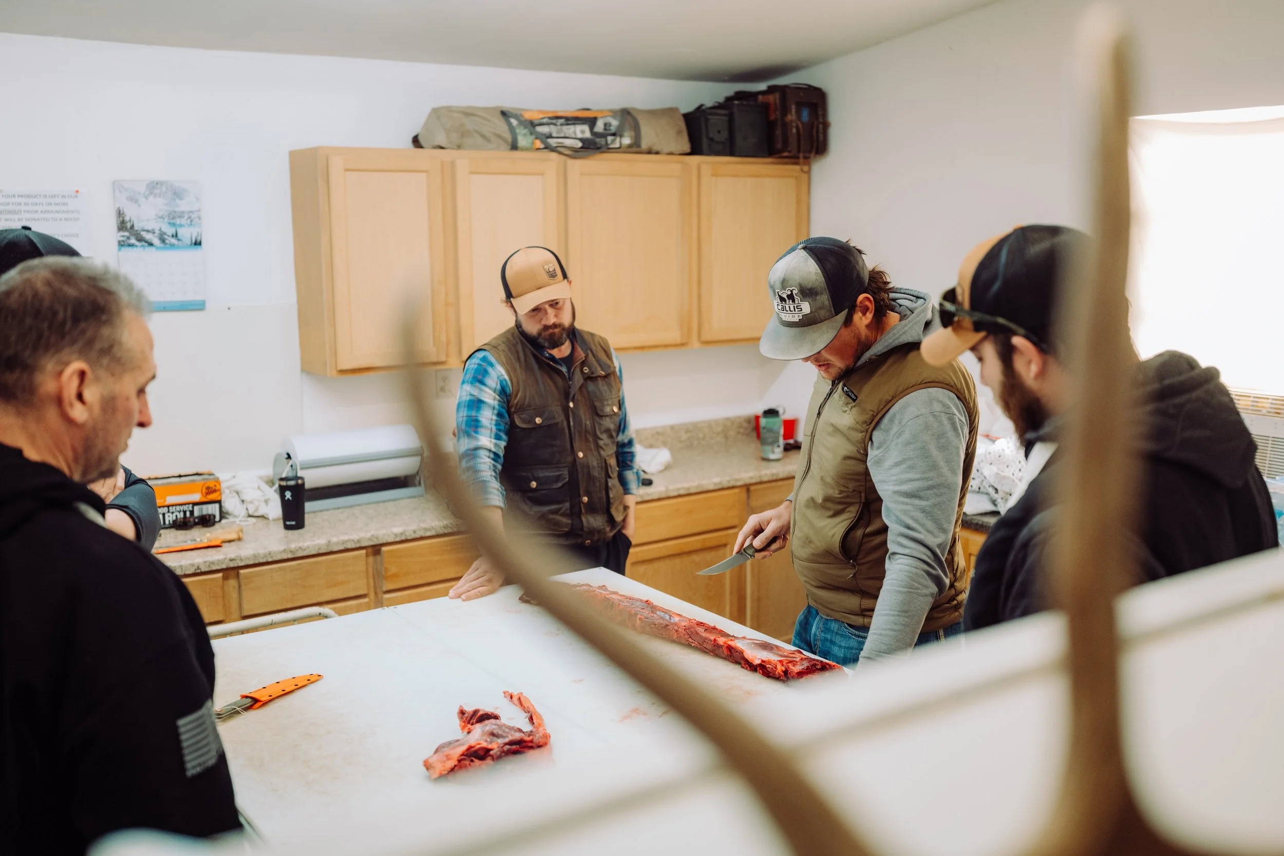 Four men in a kitchen observing a meat preparation with knives on a large white table.
