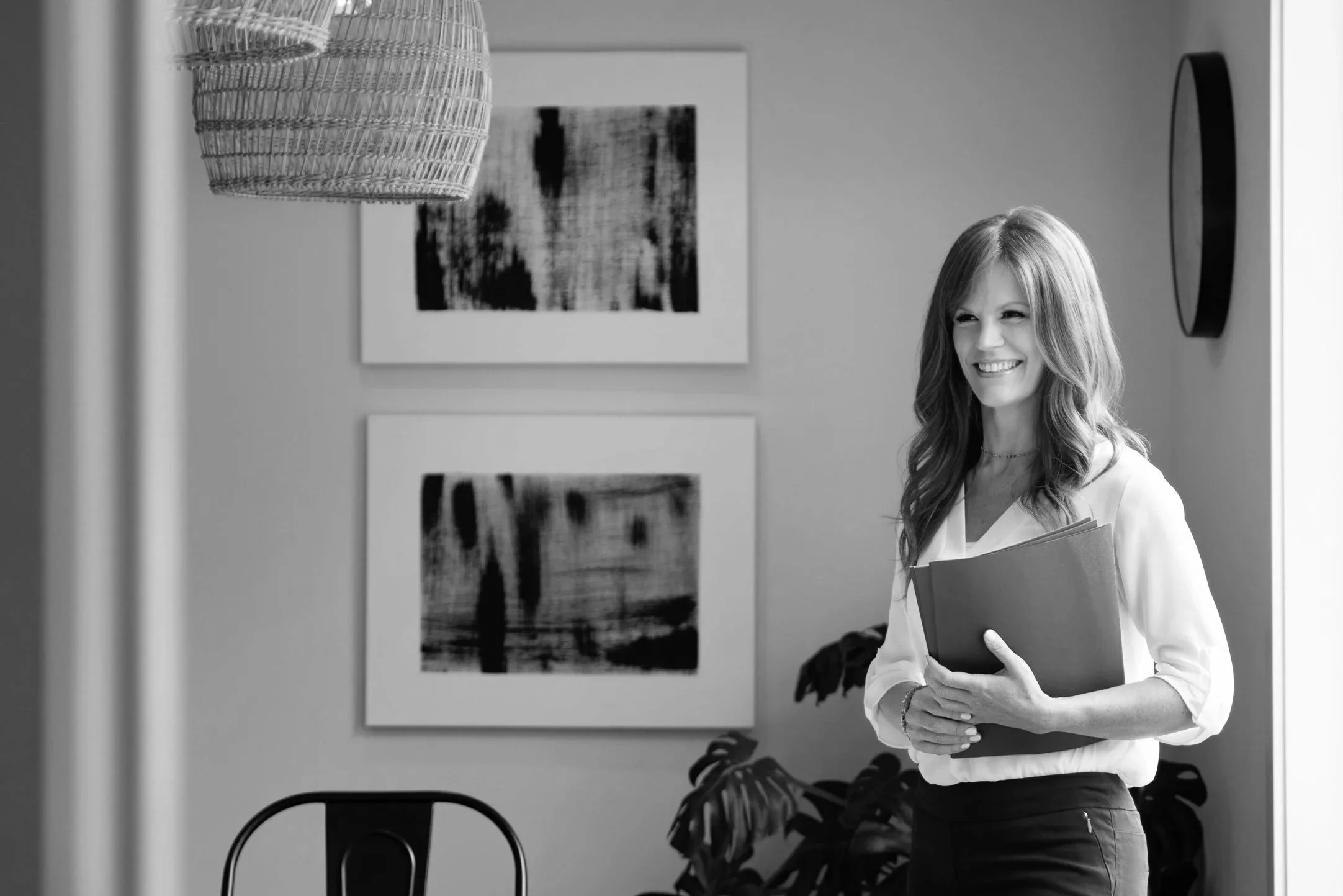 A woman with long hair smiling, holding a folder, inside a modern room with artwork and plants.