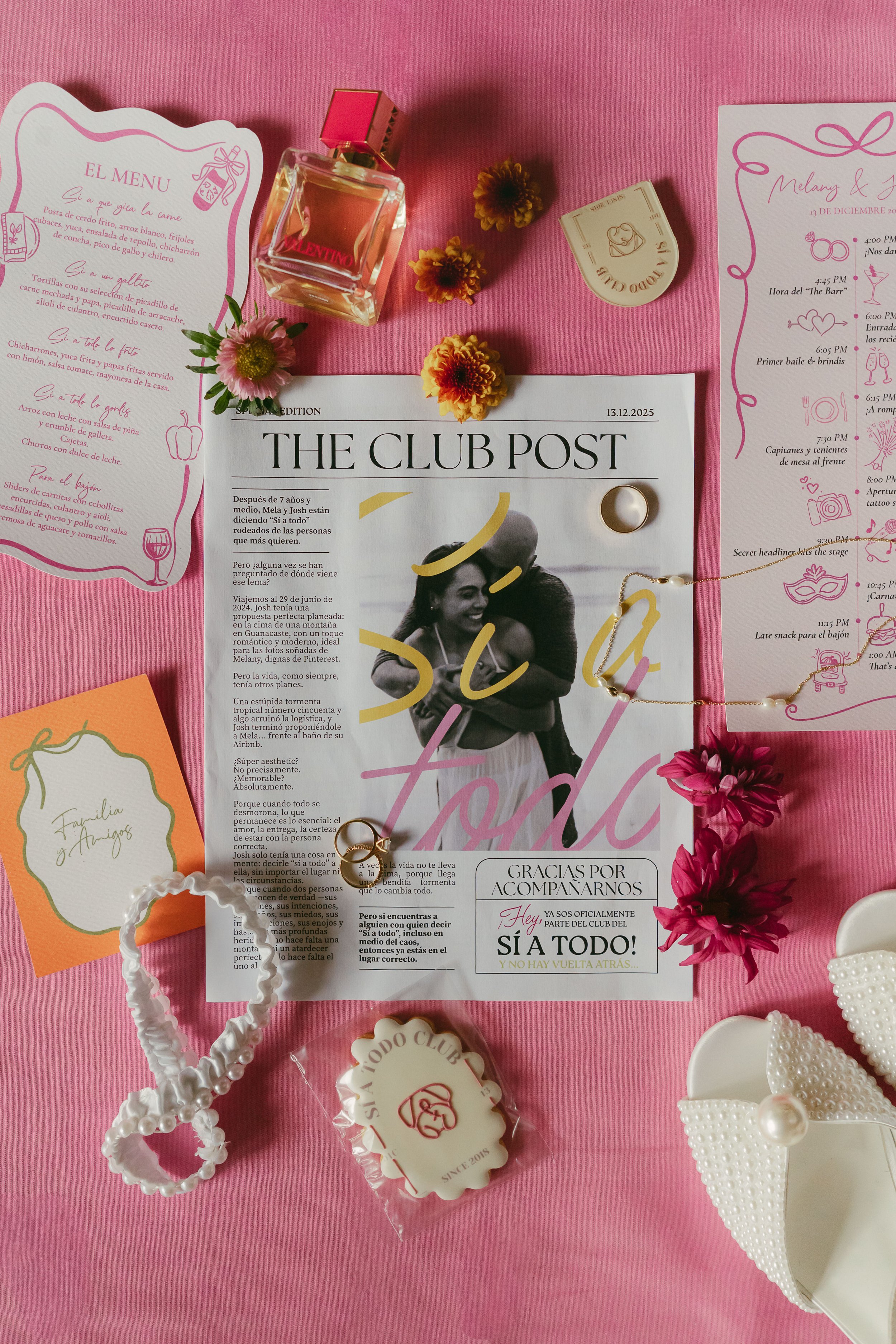 A flat lay of a pink-themed event setup with flowers, perfume, jewelry, a printed schedule, and other decorative items, including a magazine titled 'The Club Post' and a handwritten menu in Spanish.