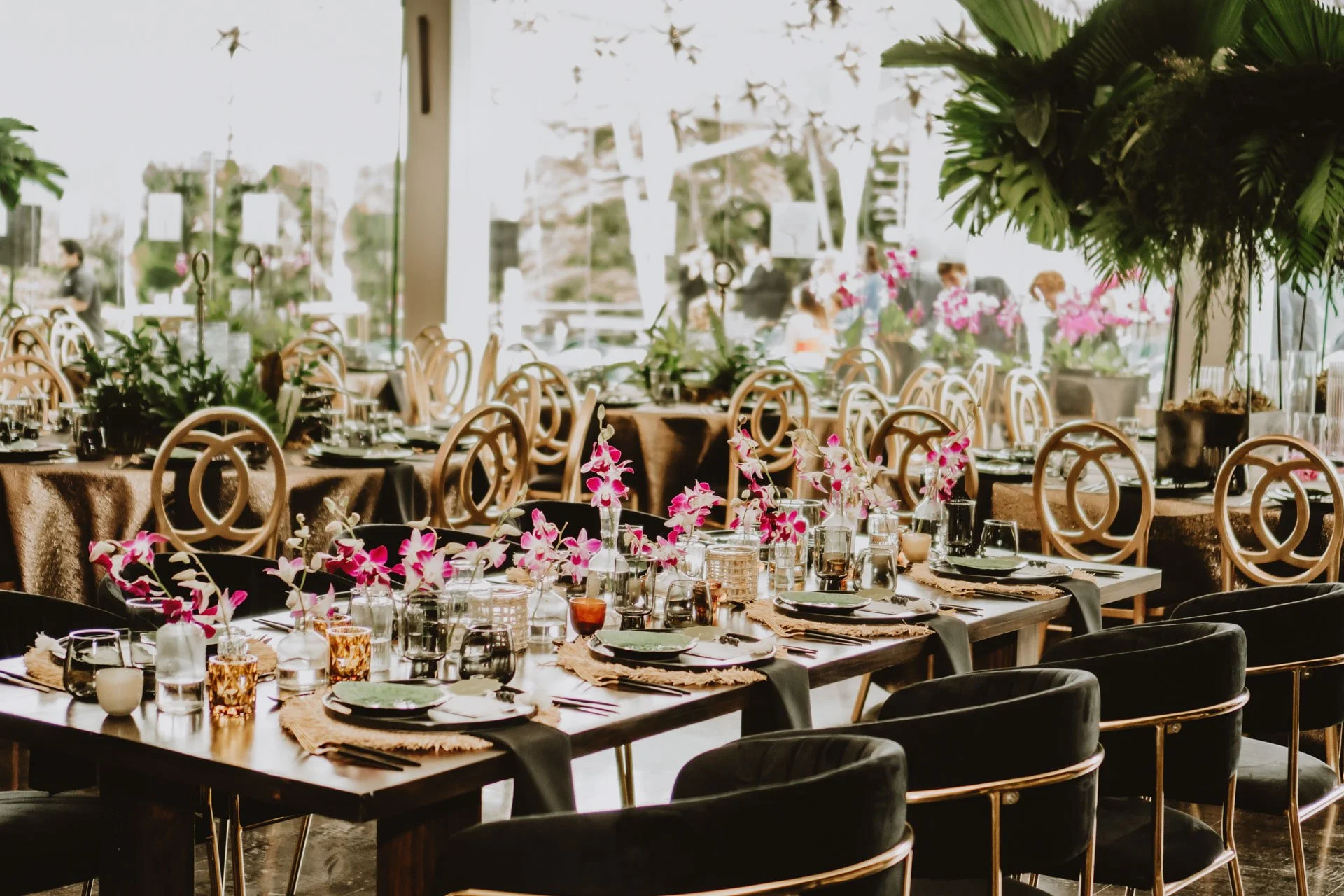 Mesas de banquete decoradas con orquídeas rosadas, cristalería y servilletas negras en un espacio interior lleno de luz natural.