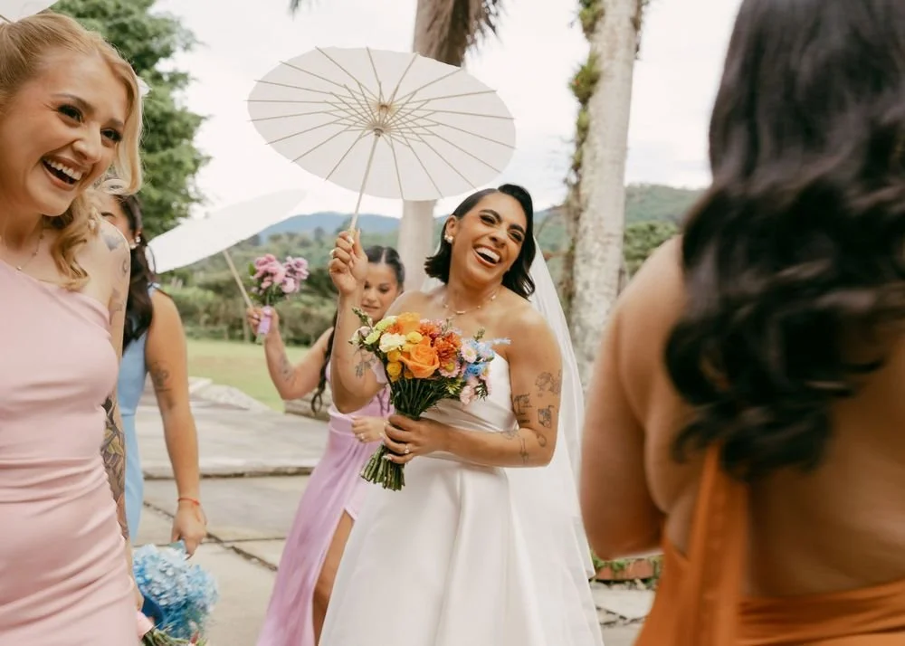 Happy bride holding a colorful bouquet at an outdoor wedding, surrounded by smiling women with umbrellas, in a scenic garden setting.