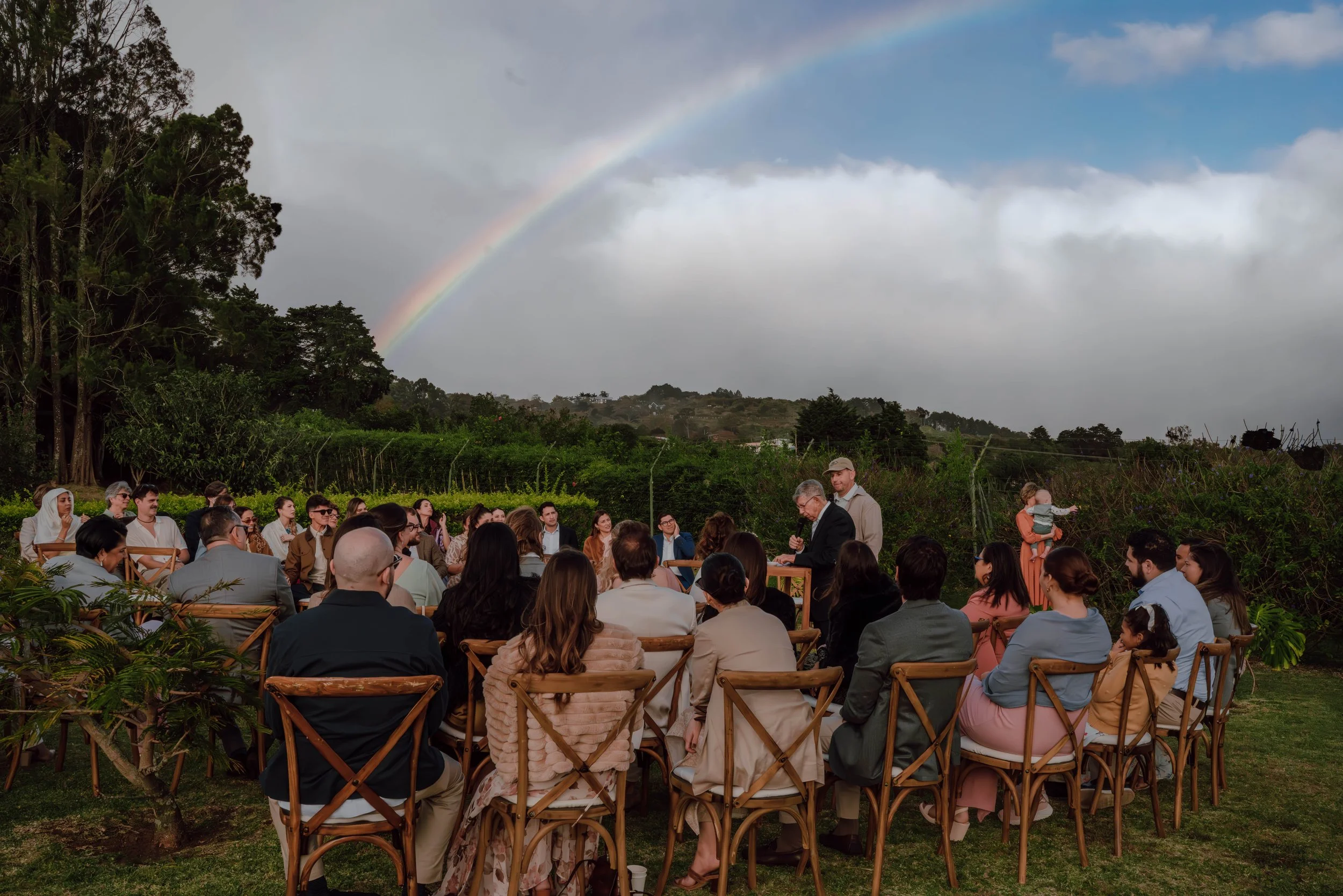 Invitados reunidos al aire libre durante una boda en Costa Rica, rodeados de árboles, vegetación y un arcoíris en el cielo.