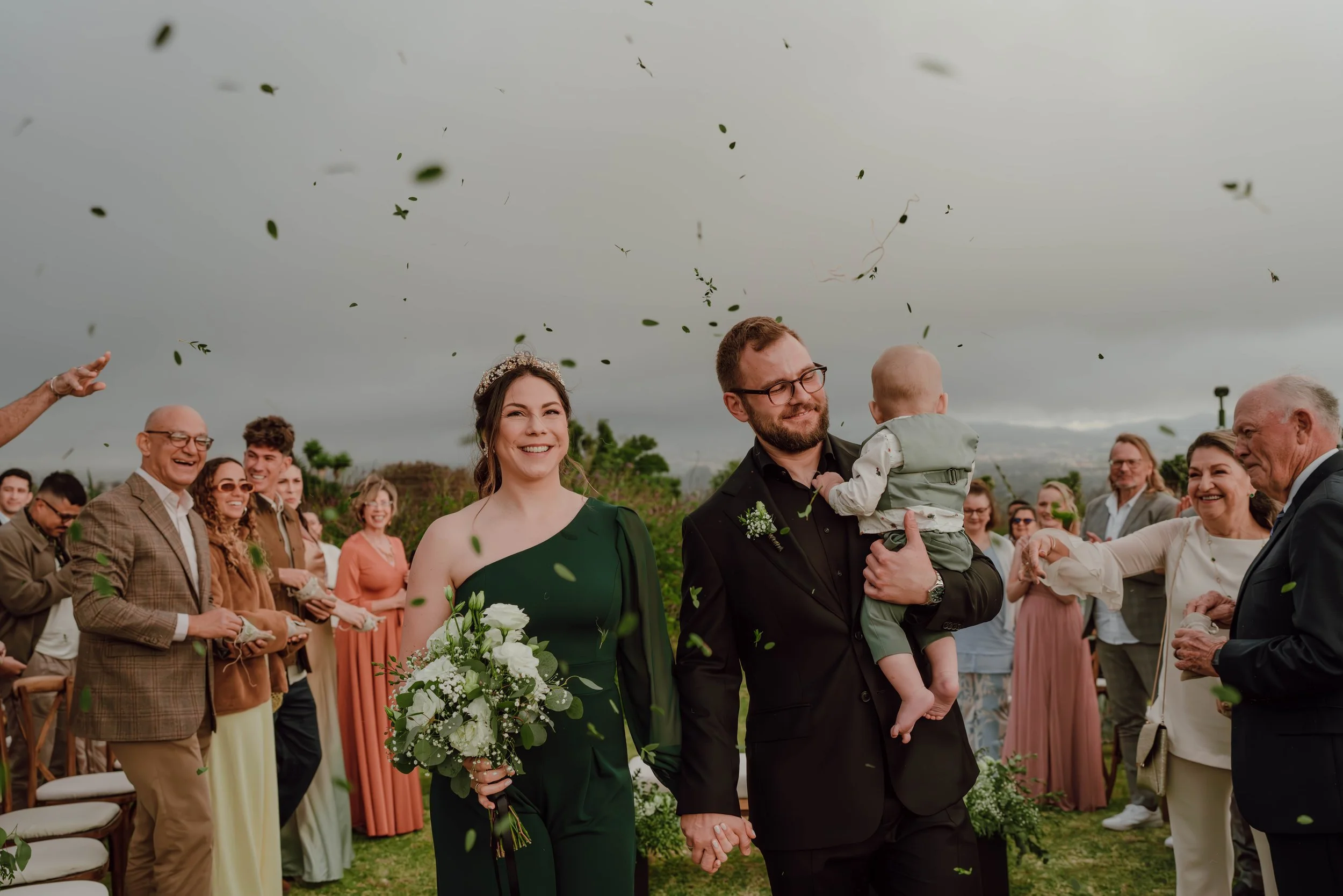 Novios tomados de la mano durante su ceremonia al aire libre, rodeados de invitados sonrientes y hojas lanzadas al aire en celebración.