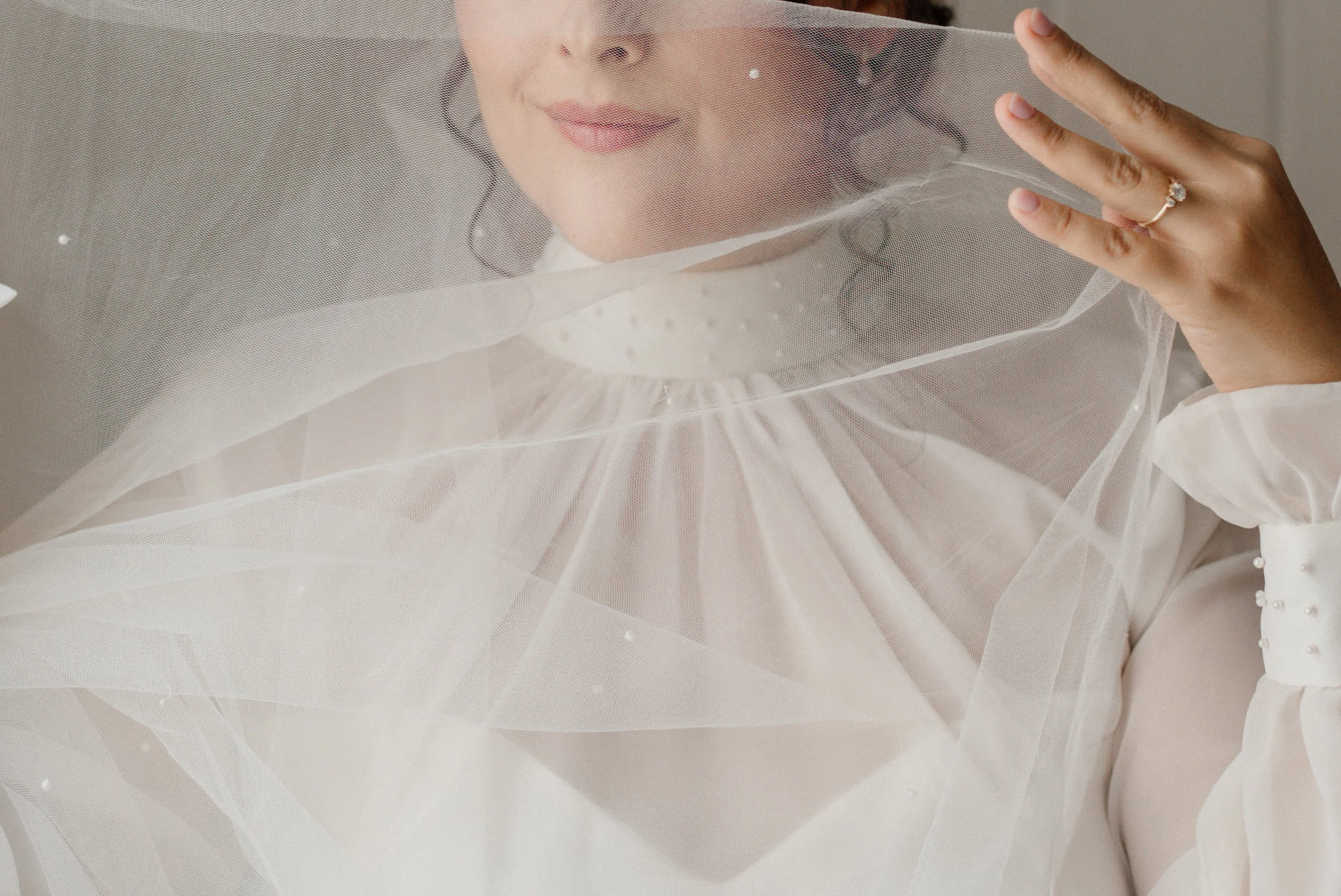 A woman in wedding attire holding a delicate veil over her face with her right hand, smiling gently.