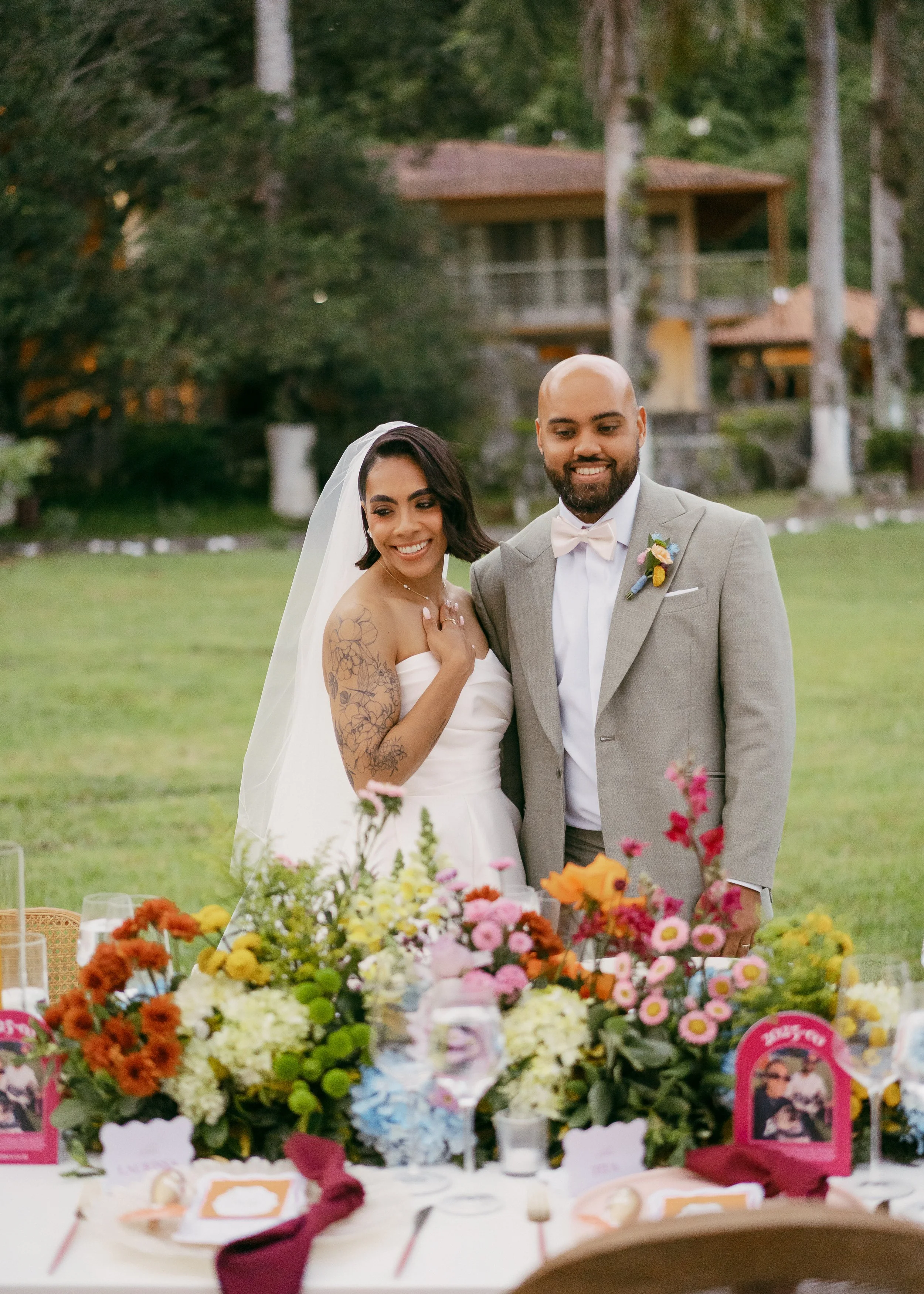A bride and groom at a wedding reception outdoors, standing behind a table decorated with colorful flowers and place settings, with a wooded background and a house or cabin.