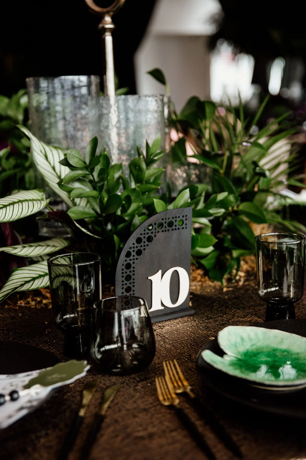 Detalle de mesa con vajilla negra, cubiertos dorados, servilletas estampadas, cristalería y número de mesa rodeado de vegetación.