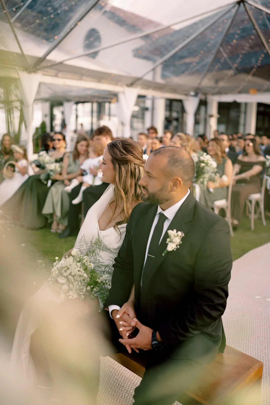 Pareja sentada durante la ceremonia de su boda, con invitados al fondo bajo una carpa.