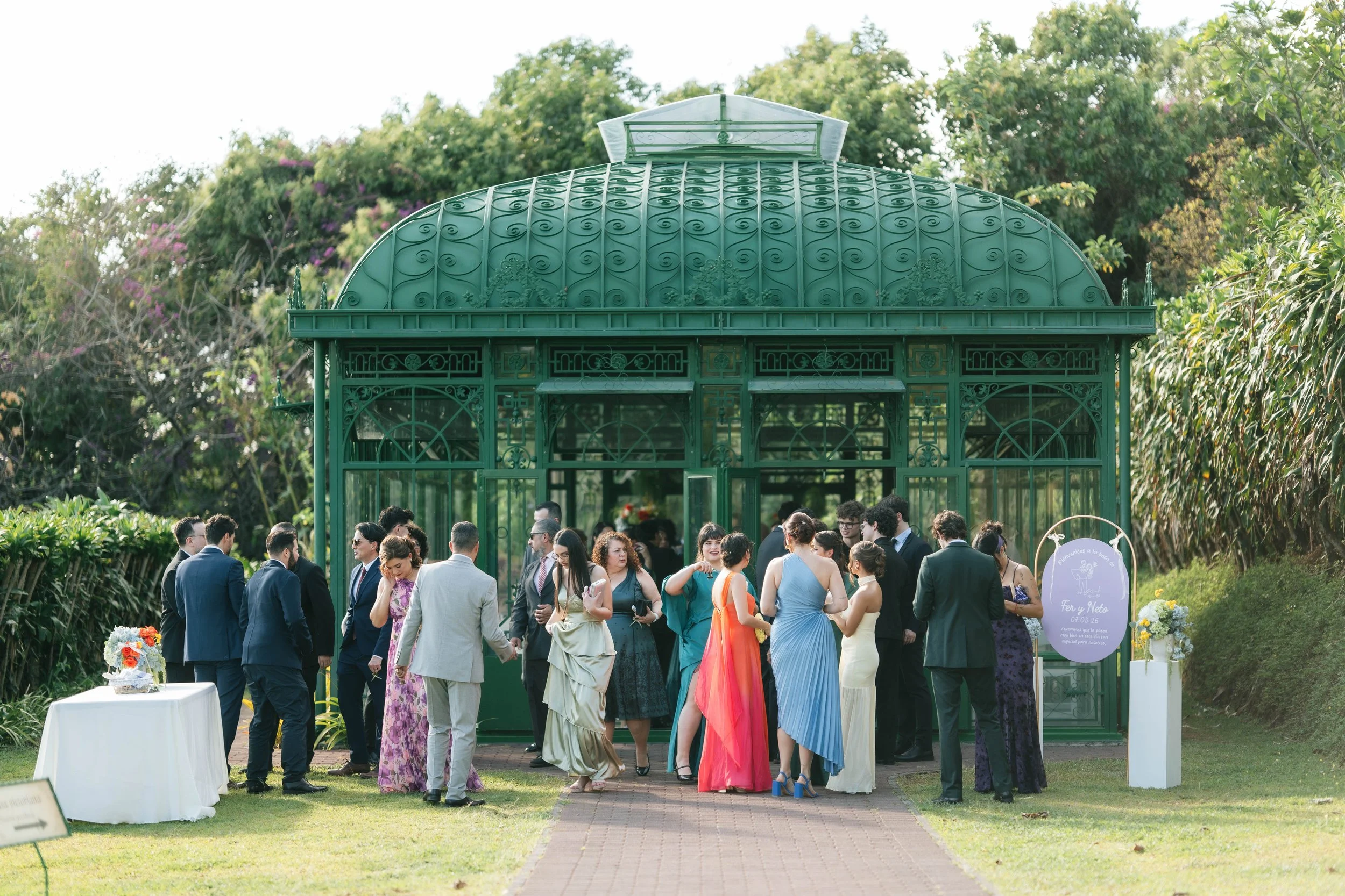 Ceremonia de boda en jardín con invitados reunidos frente a un invernadero ornamental verde.