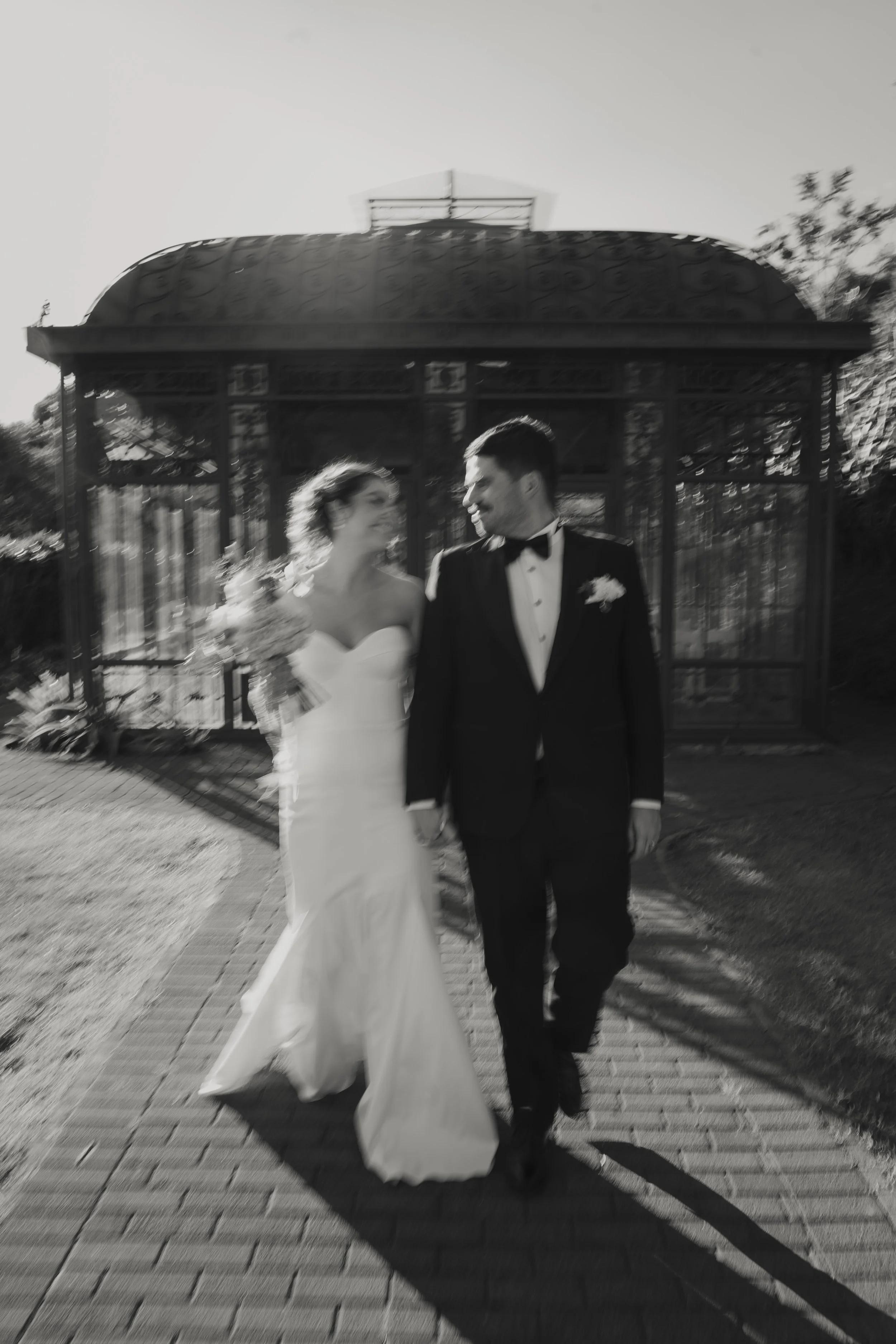 A bride and groom walking together outdoors, smiling at each other, with a metal gazebo and trees in the background, in an outdoor wedding setting.