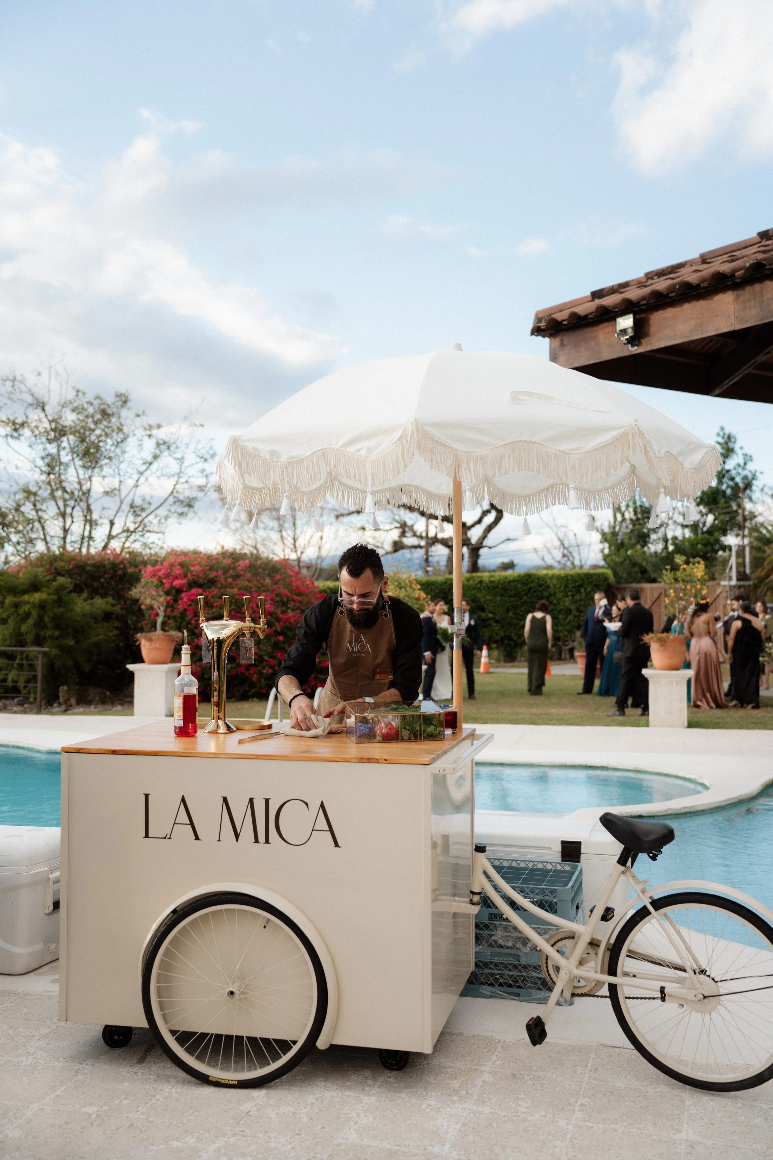 Persona preparando bebidas en una barra móvil tipo bicicleta durante una boda al aire libre, con piscina y vegetación alrededor.