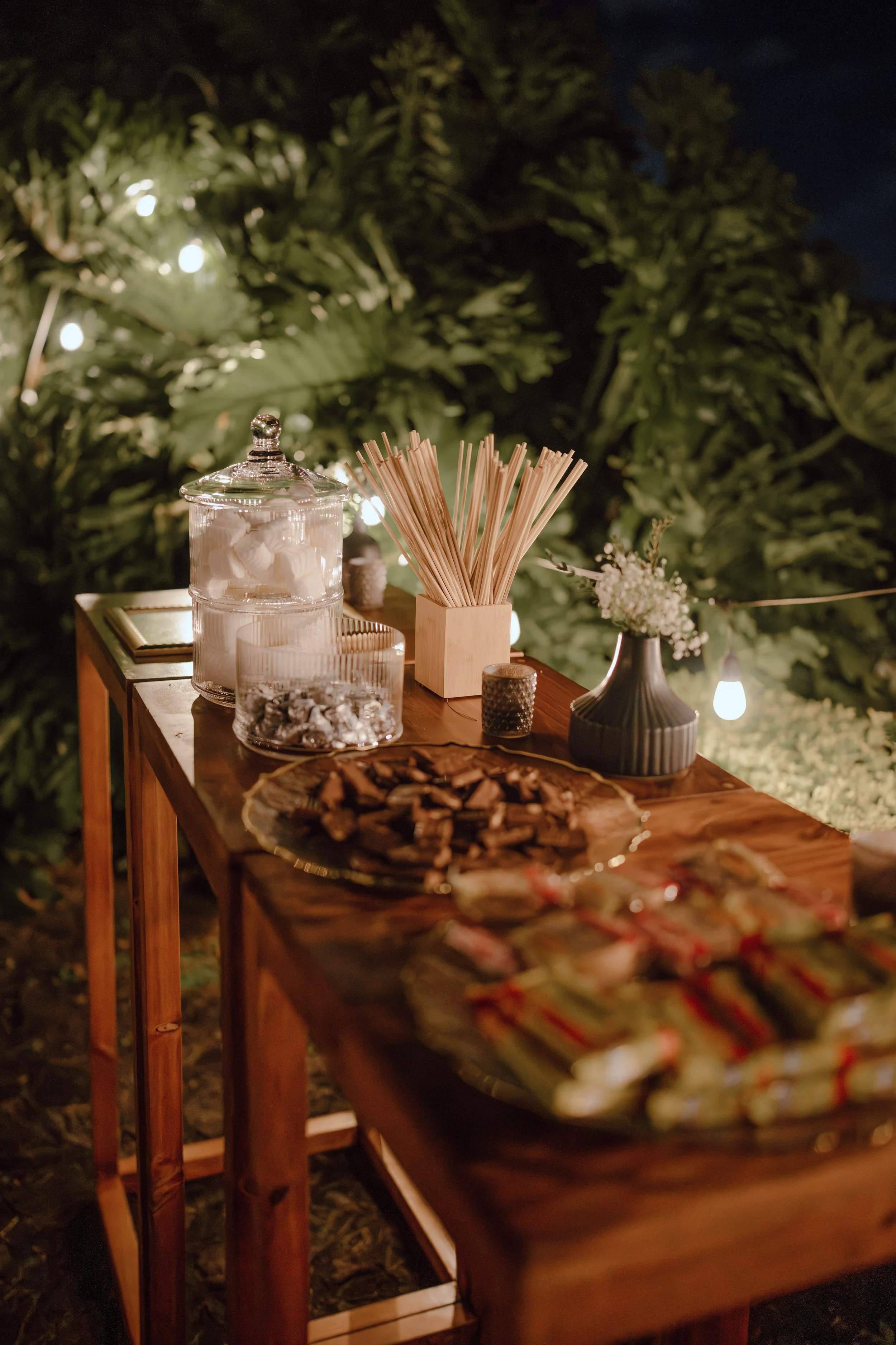 Mesa de estación decorada con snacks, frascos de dulces, flores blancas y luces cálidas durante una boda al aire libre en Costa Rica.