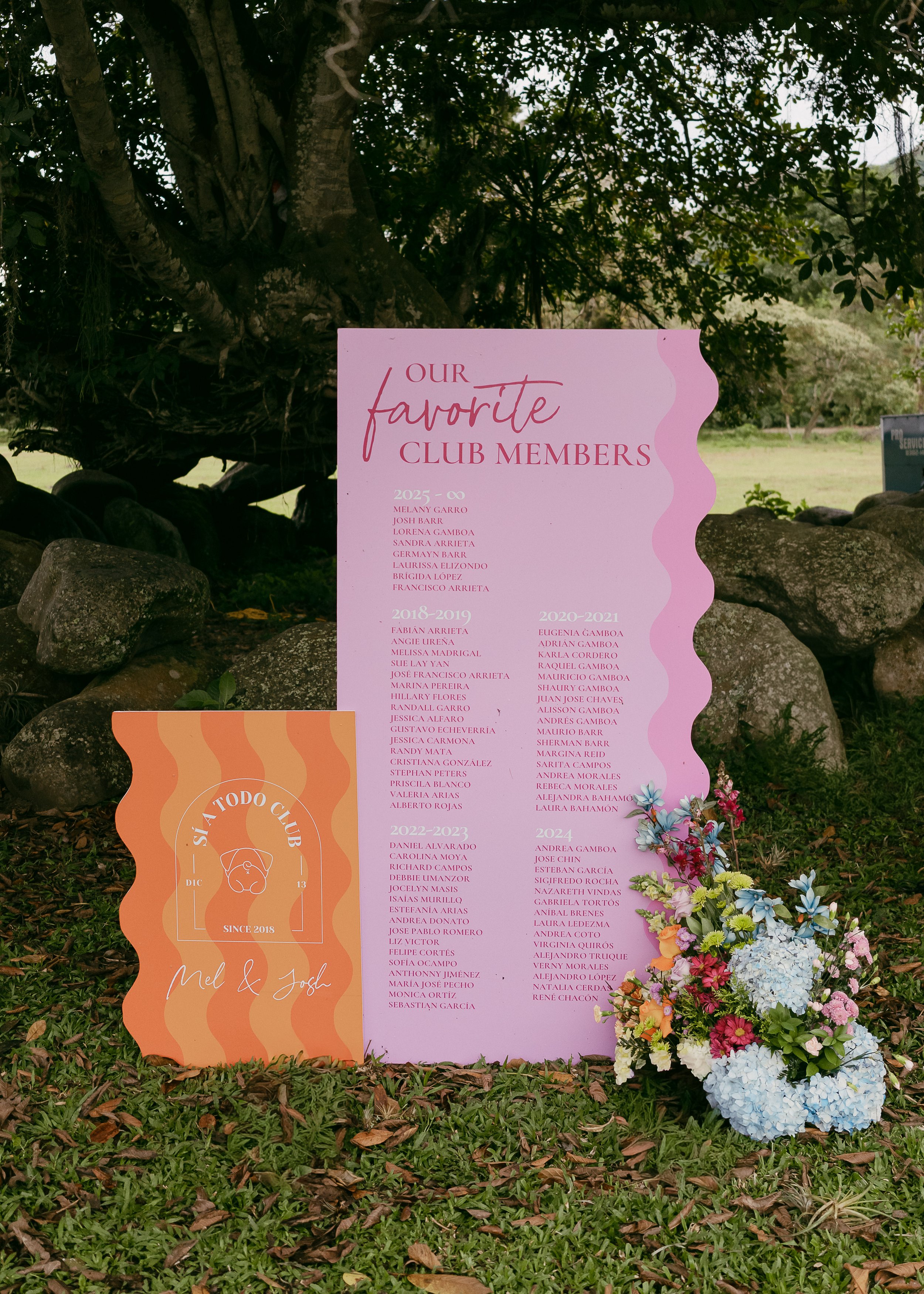 Signboard listing favorite club members from 2018 - 2025, with a decorative orange section at the bottom featuring the names Mel and Josh and a floral arrangement at the bottom right corner. The sign is outdoors, surrounded by rocks and grass.