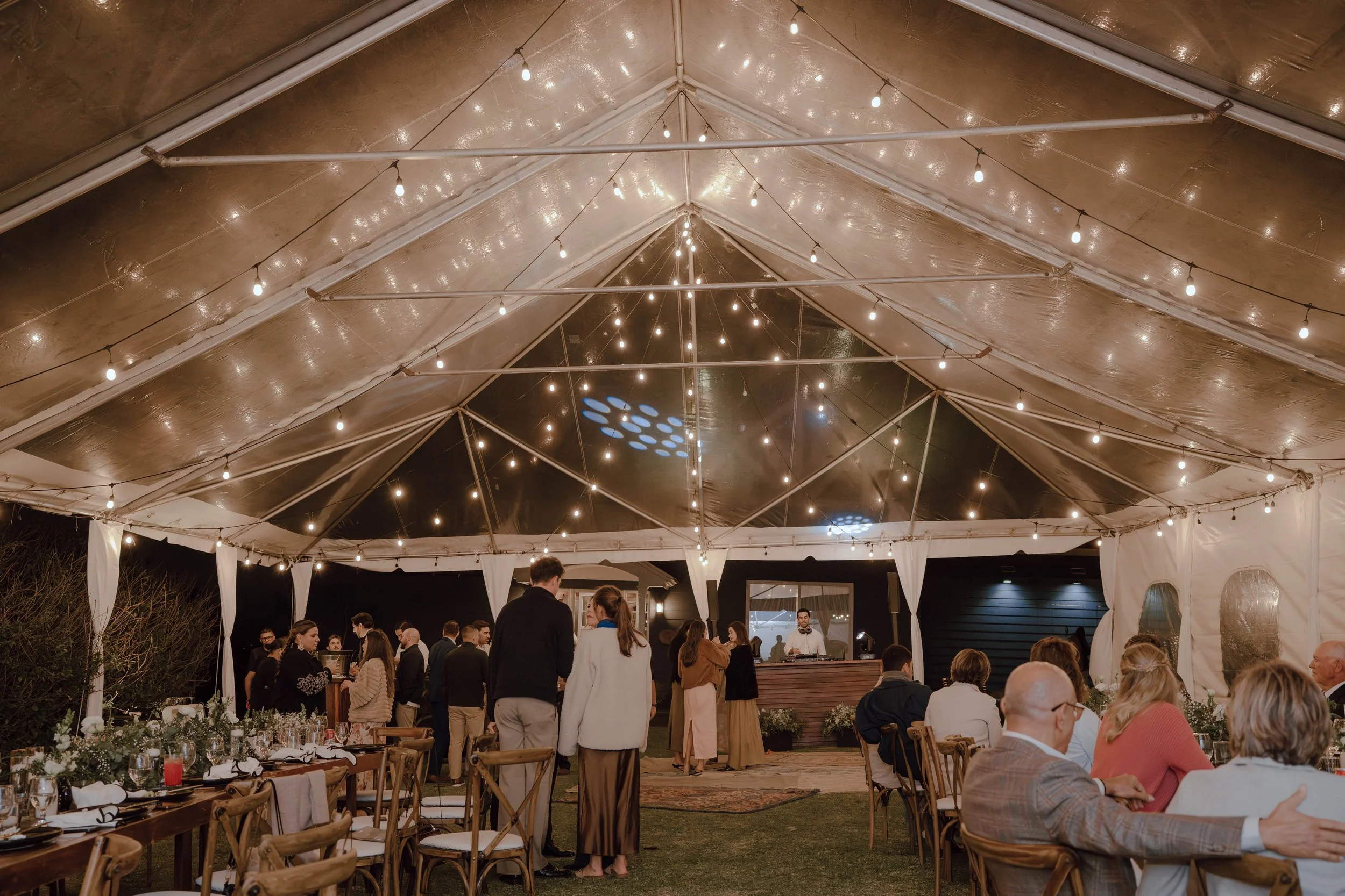 Invitados socializando bajo una carpa con luces cálidas y mesas preparadas para la recepción de una boda al aire libre.