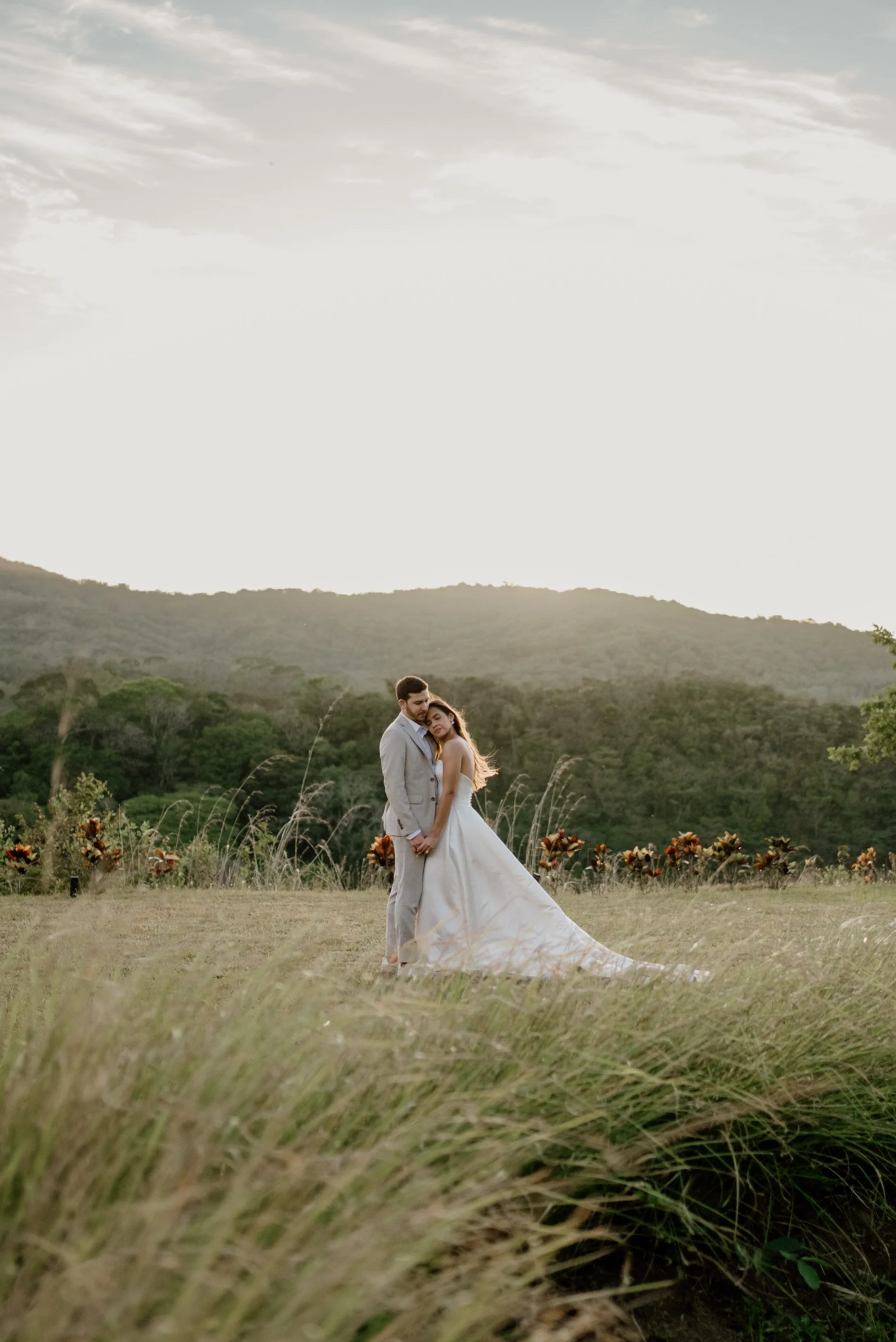 A couple dressed in wedding attire standing in a grassy field with mountains and trees in the background, during sunset.