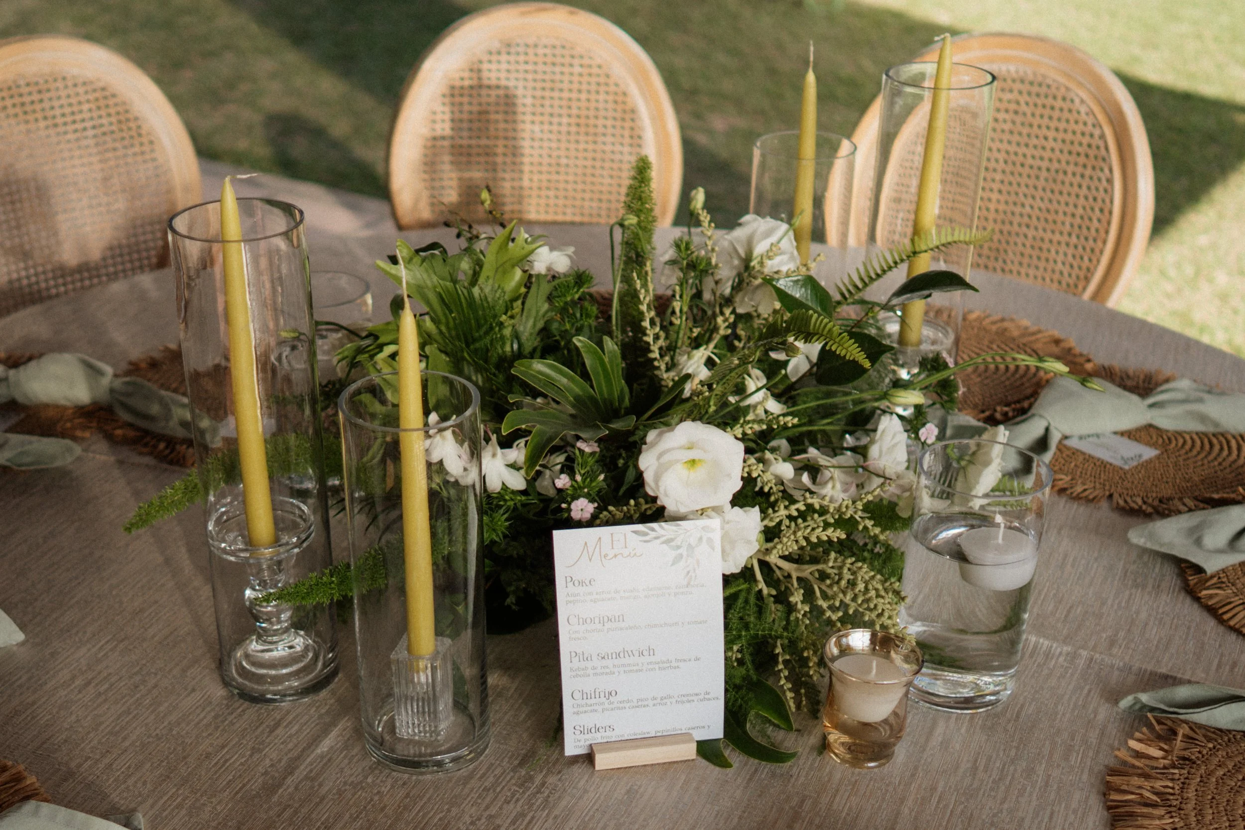 Detalle de mesa al aire libre con centro floral, velas amarillas, menú y sillas de fibras naturales durante una boda en Costa Rica.