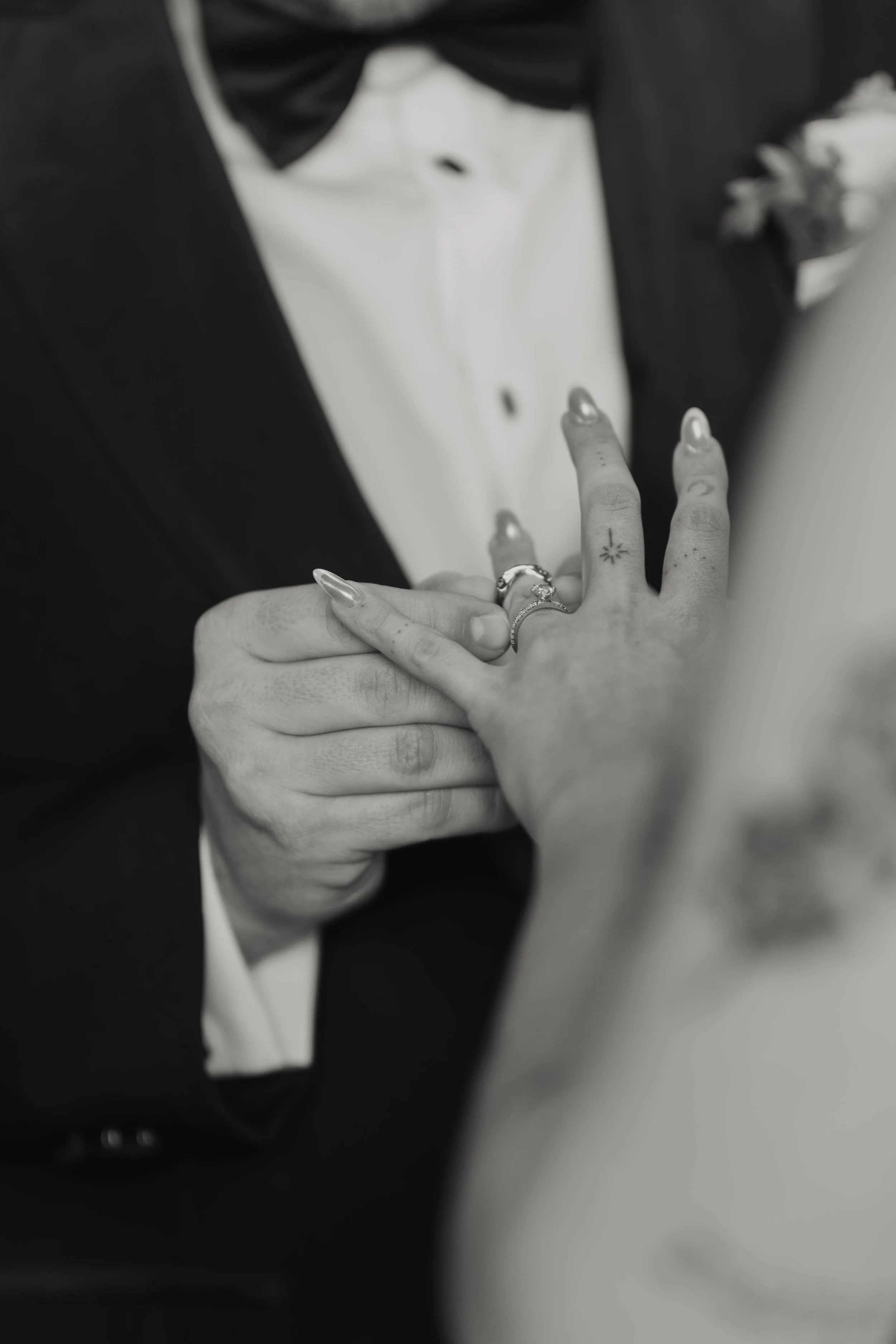 Fotografía en blanco y negro de las manos de los novios, con anillos y vestimenta formal durante su boda.