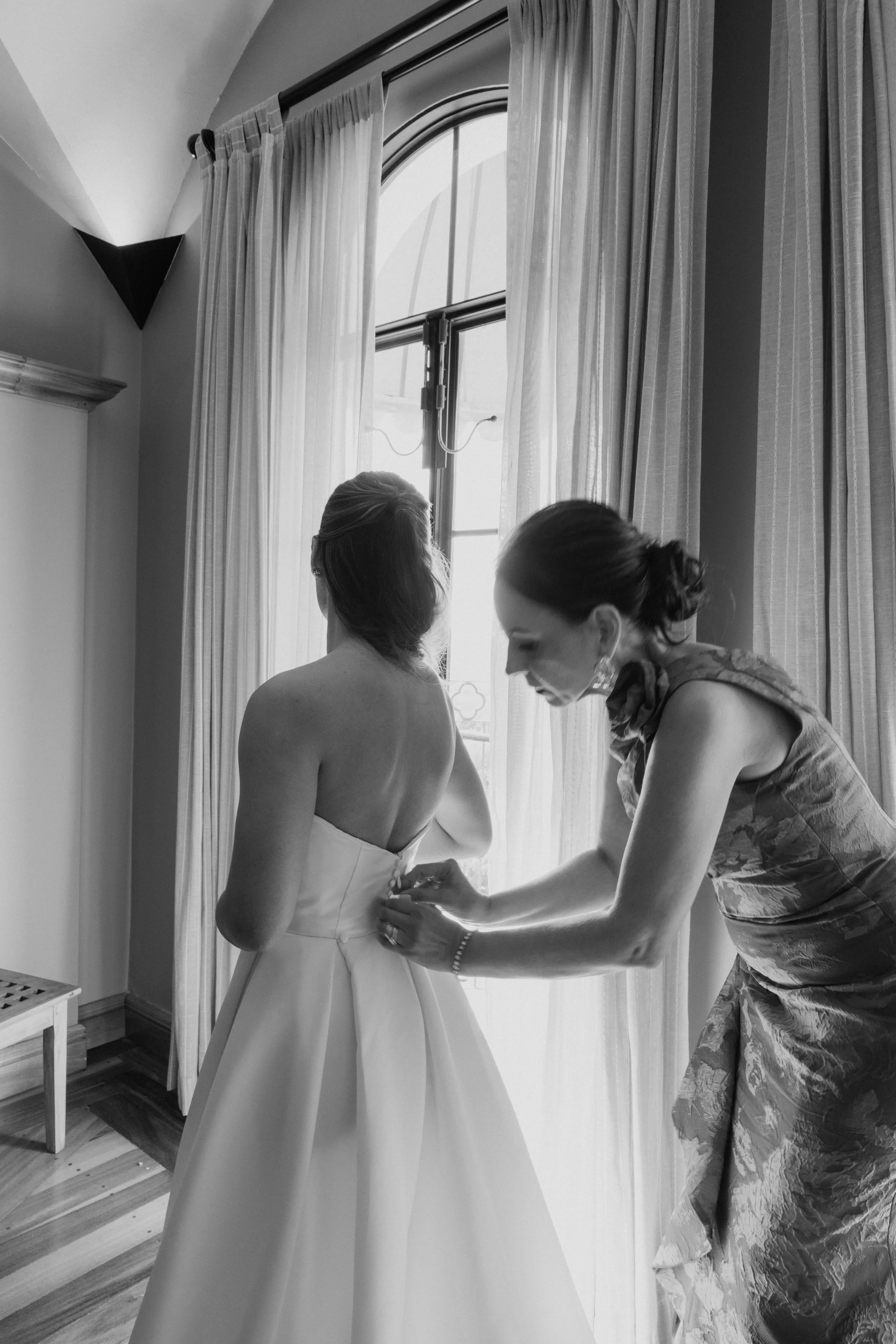 A bride in a strapless wedding gown is having her dress buttoned by her mother in an elegant dress, as they stand near a large window with sheer curtains.