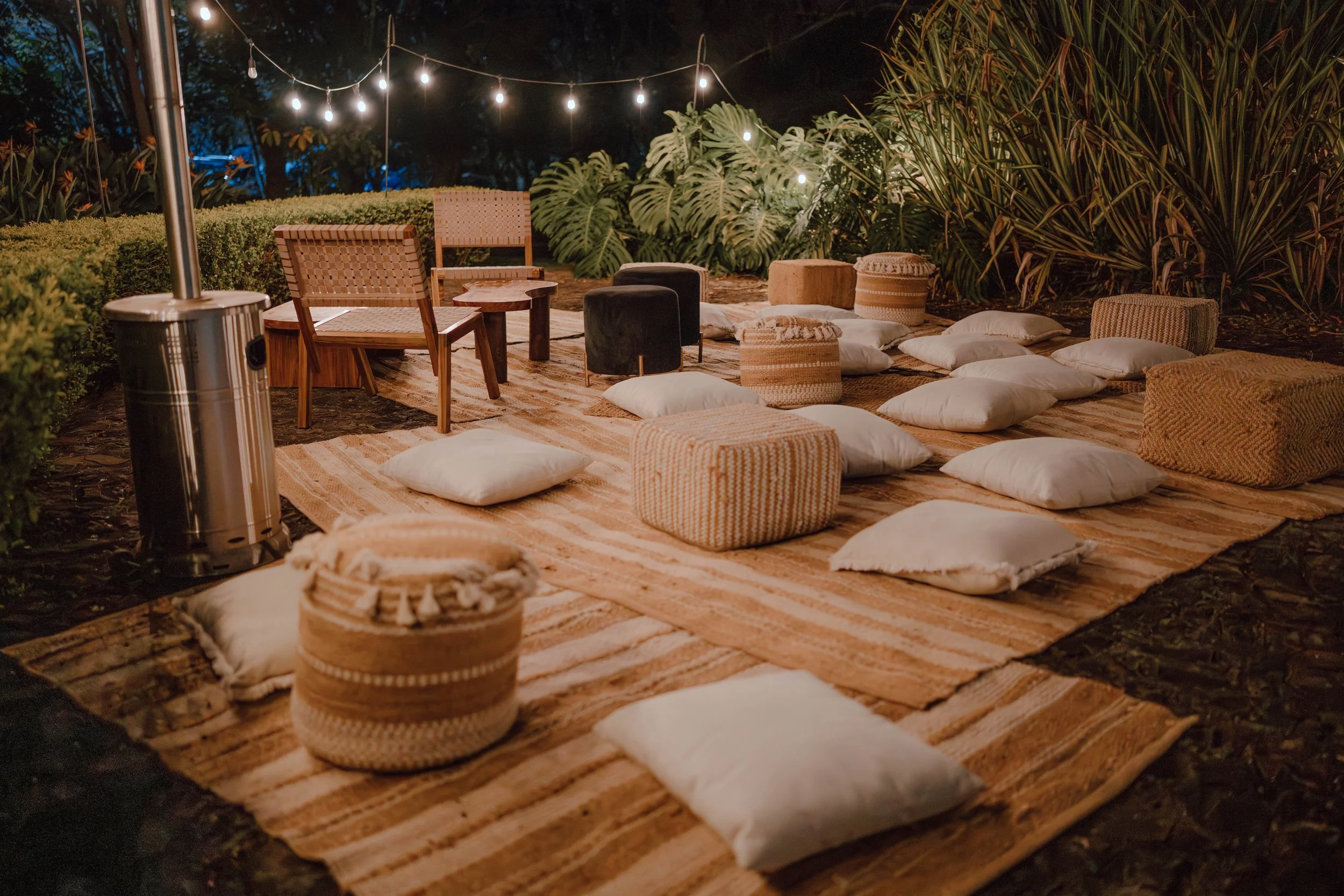 Espacio de descanso al aire libre durante una boda nocturna, con guirnaldas de luces, sillas de mimbre, cojines y vegetación alrededor.