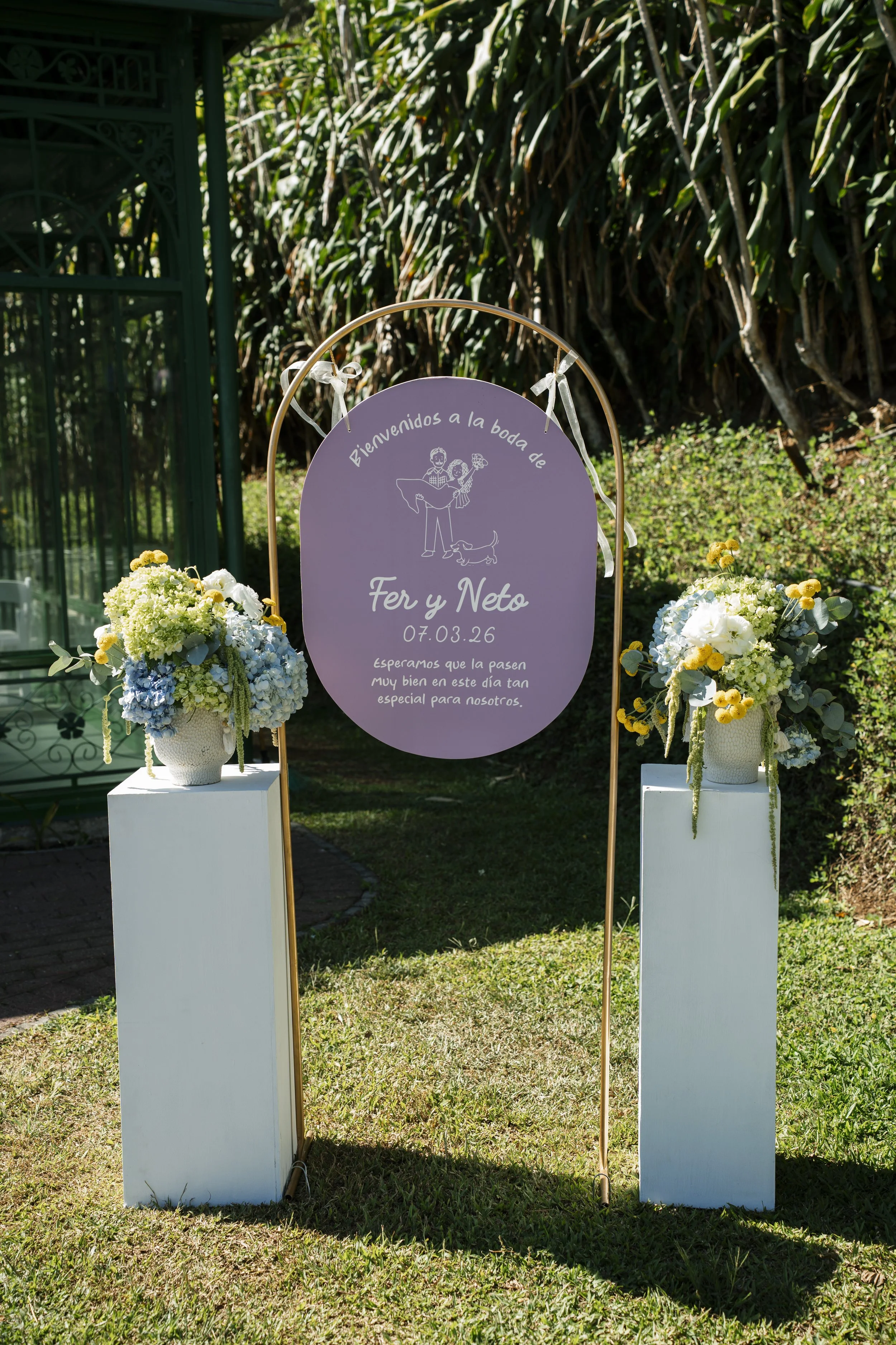 Señalización decorativa para una boda al aire libre, con texto blanco sobre fondo morado y flores en bases blancas.