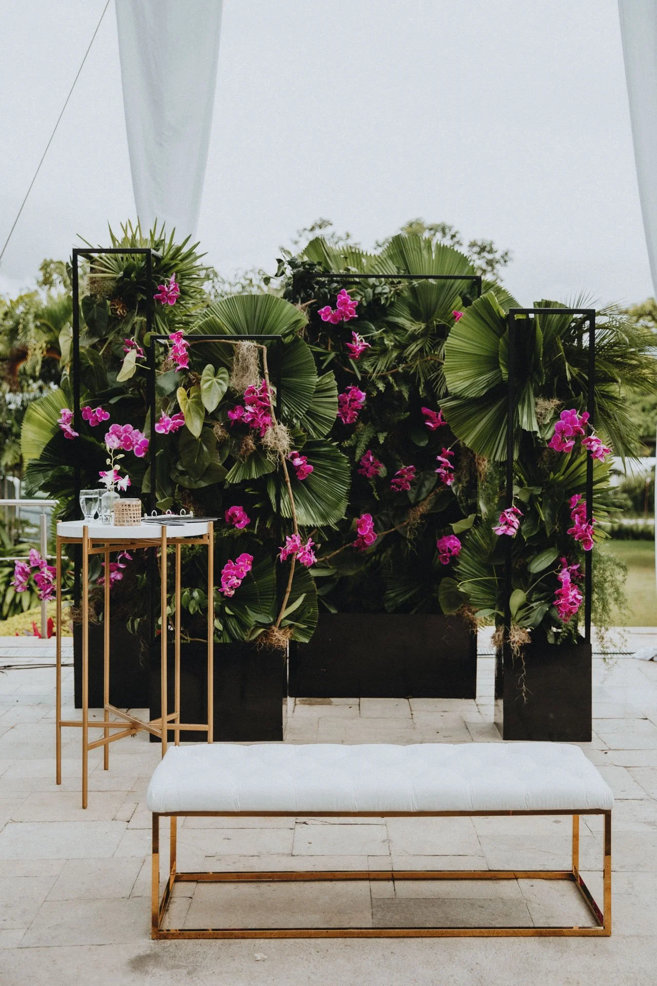 Back decorativo con orquídeas rosadas, hojas de palma, marcos negros y área lounge al aire libre durante una boda en Costa Rica.