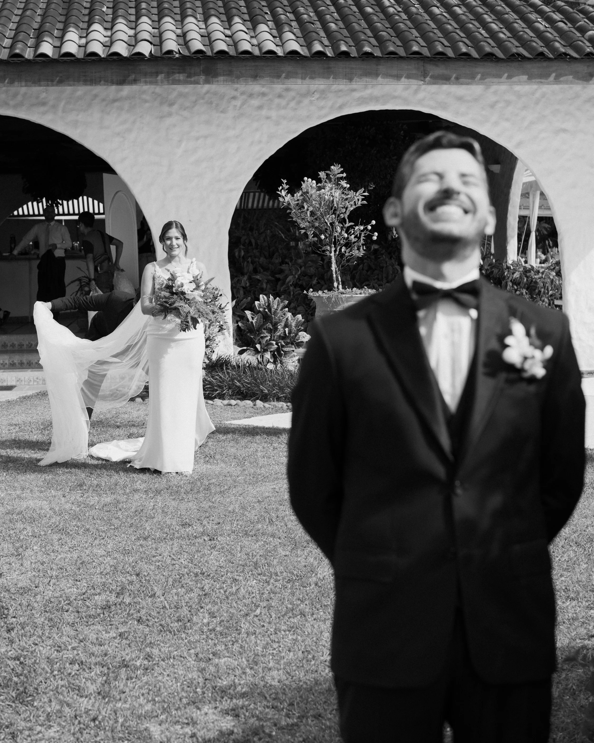 Novia con bouquet bajo un arco en una fotografía de boda en blanco y negro, con el novio desenfocado en primer plano.