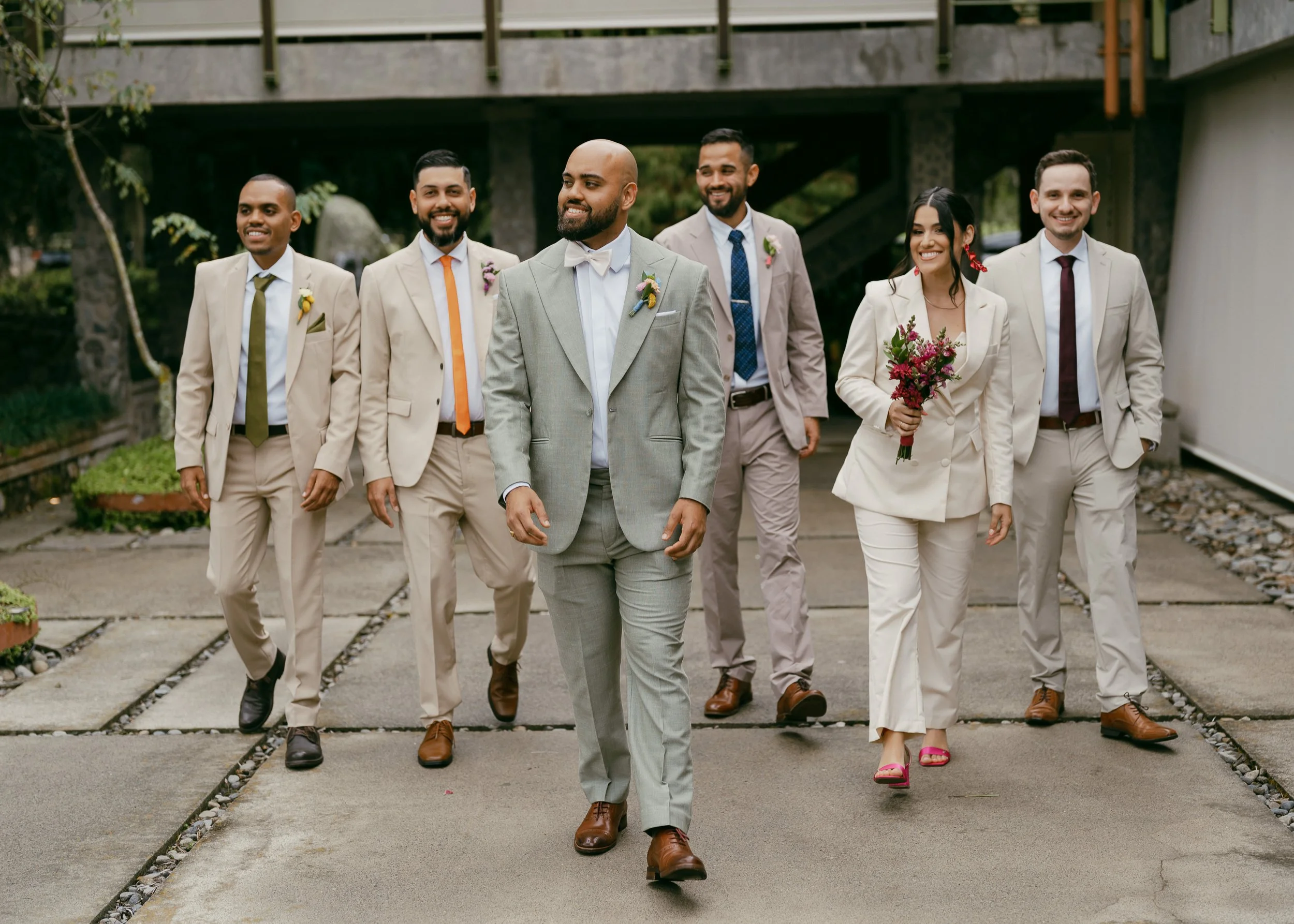 Group of people walking outdoors in wedding attire, including a person holding a bouquet of flowers.