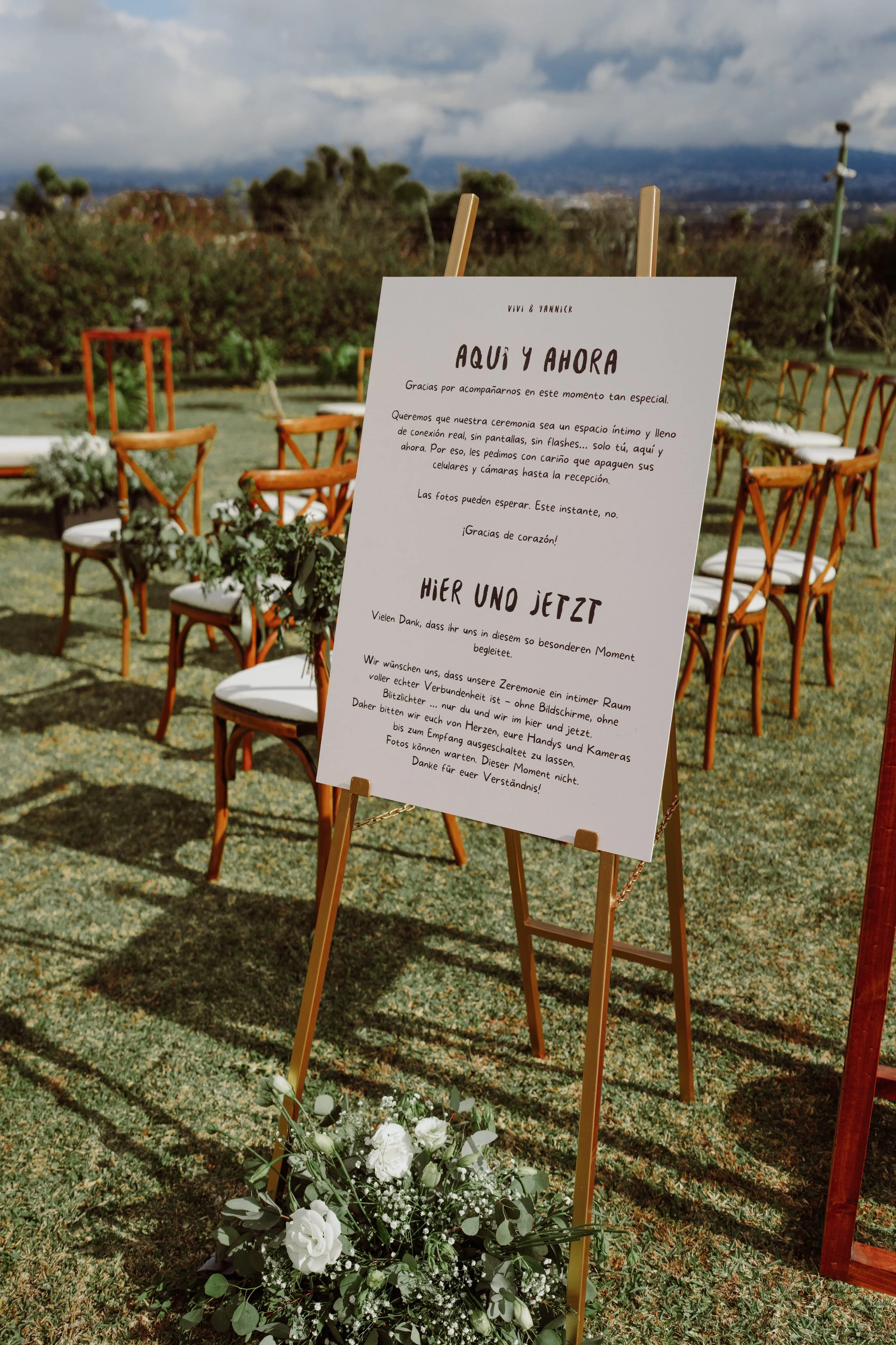 Espacio de boda al aire libre con sillas alineadas, decoración floral y letrero de bienvenida, rodeado de paisaje natural y cielo nublado.