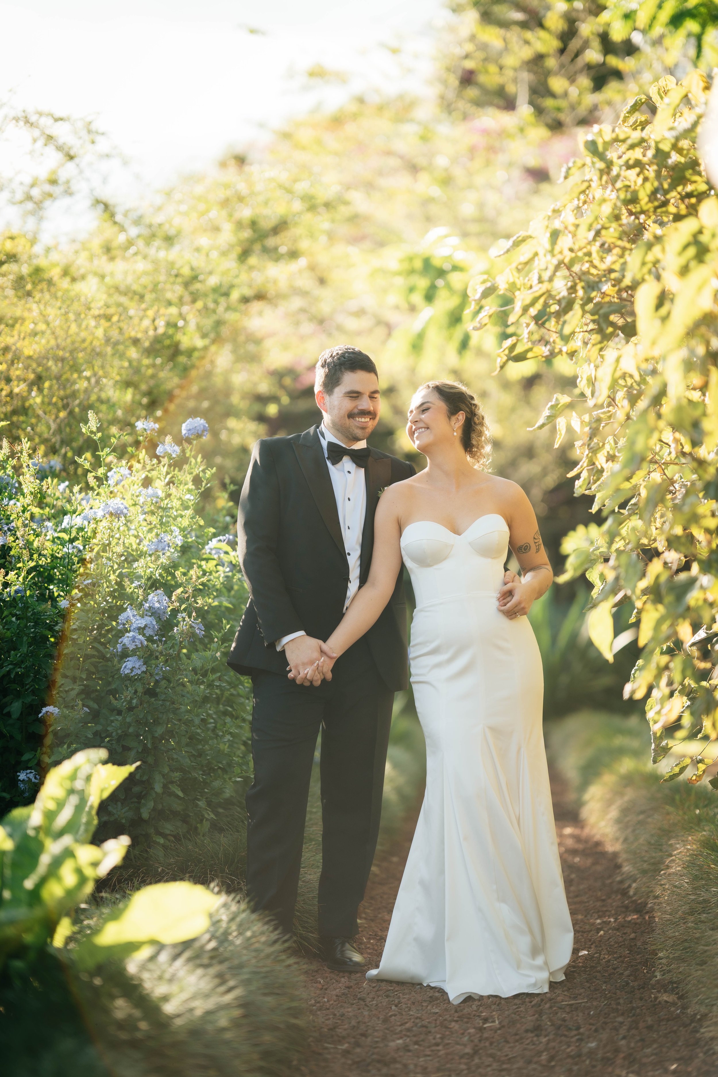 A newlywed couple walking hand in hand through a lush garden path, smiling at each other in the sunlight.