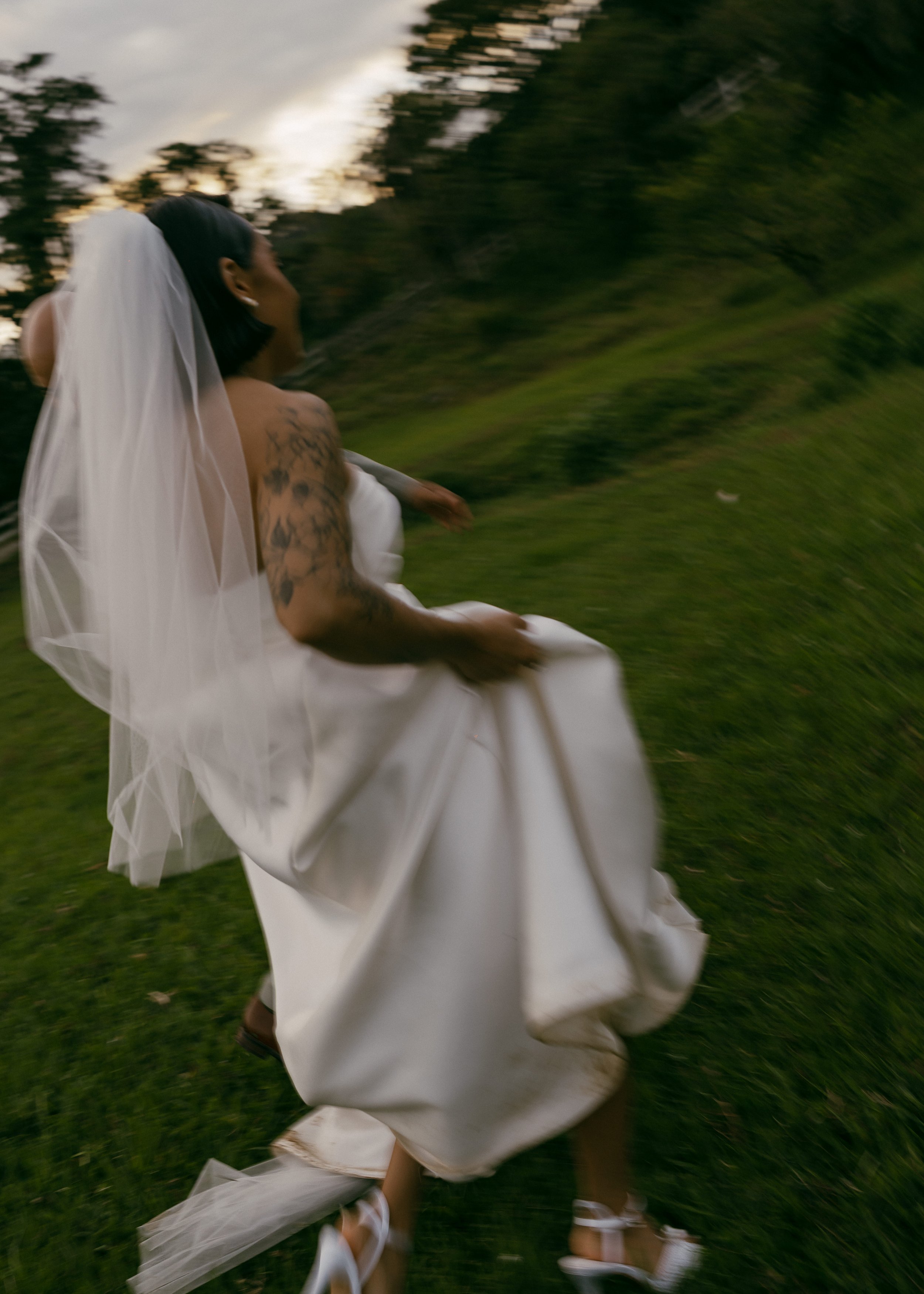 A woman in a wedding dress and veil running or moving quickly outdoors on grass with trees in the background during sunset.