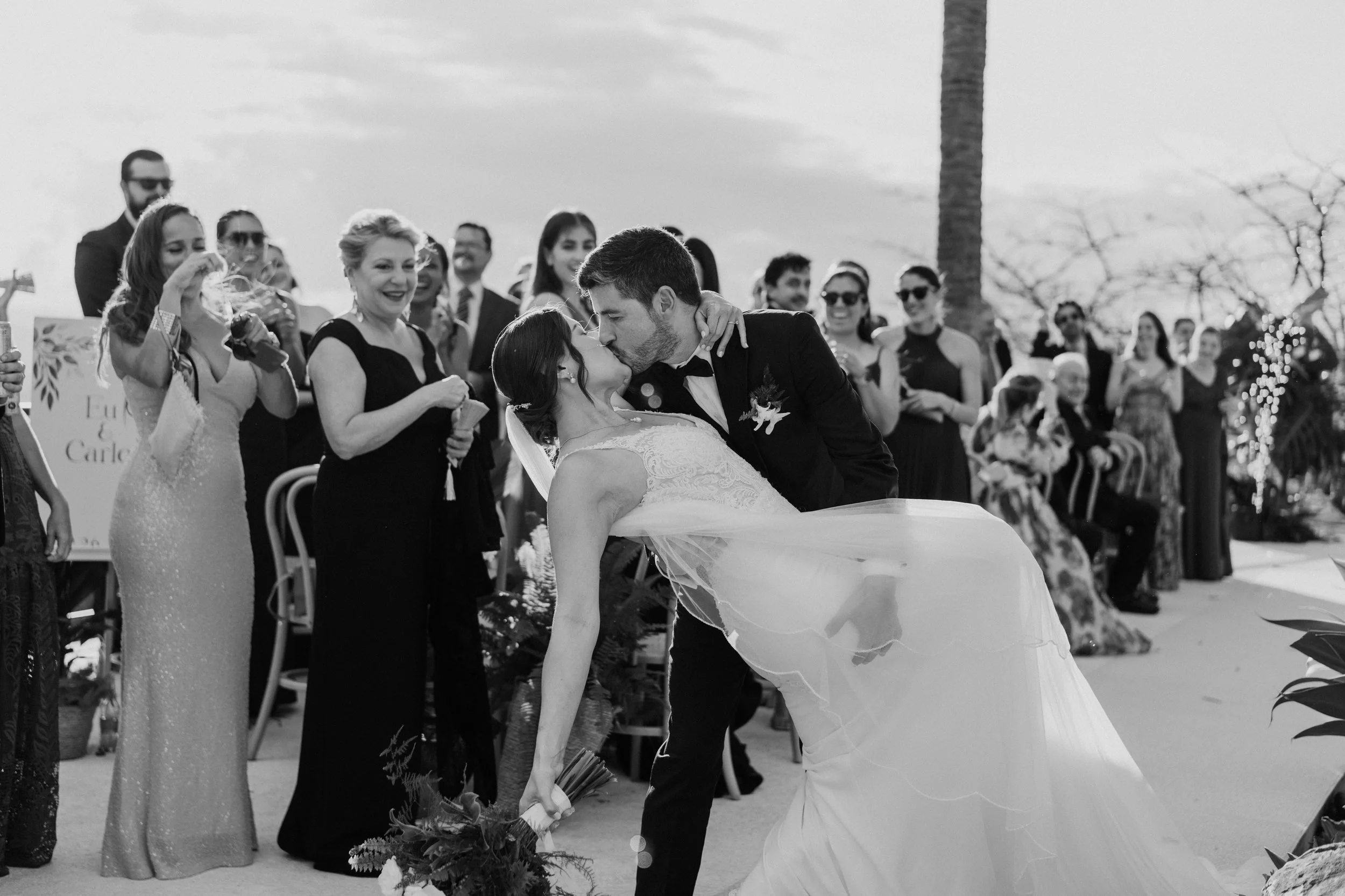Beso de los novios durante su boda en una fotografía en blanco y negro, mientras los invitados observan con alegría.