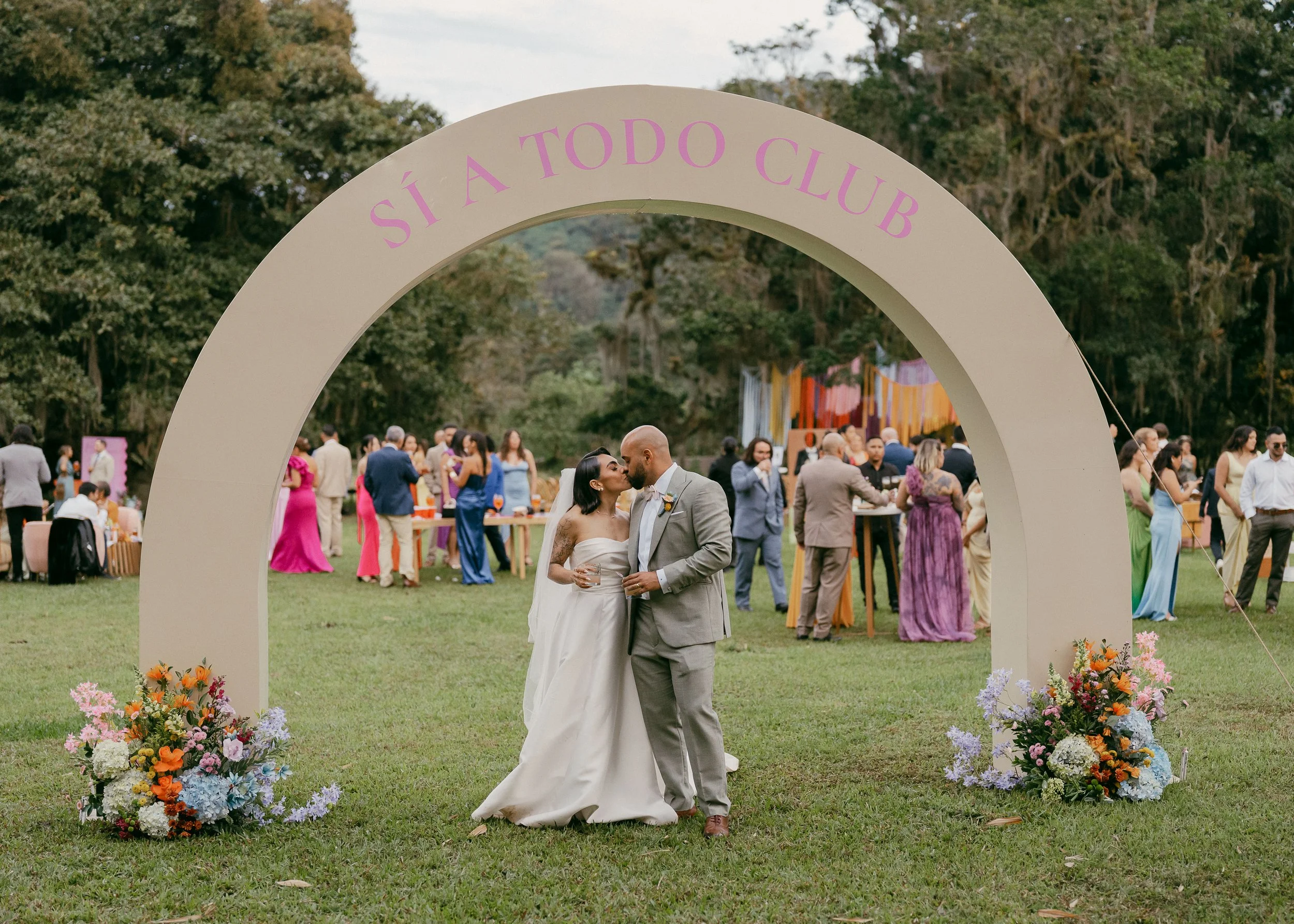 A bride and groom standing under an arch at their outdoor wedding reception, with guests in colorful attire in the background and trees surrounding the area.