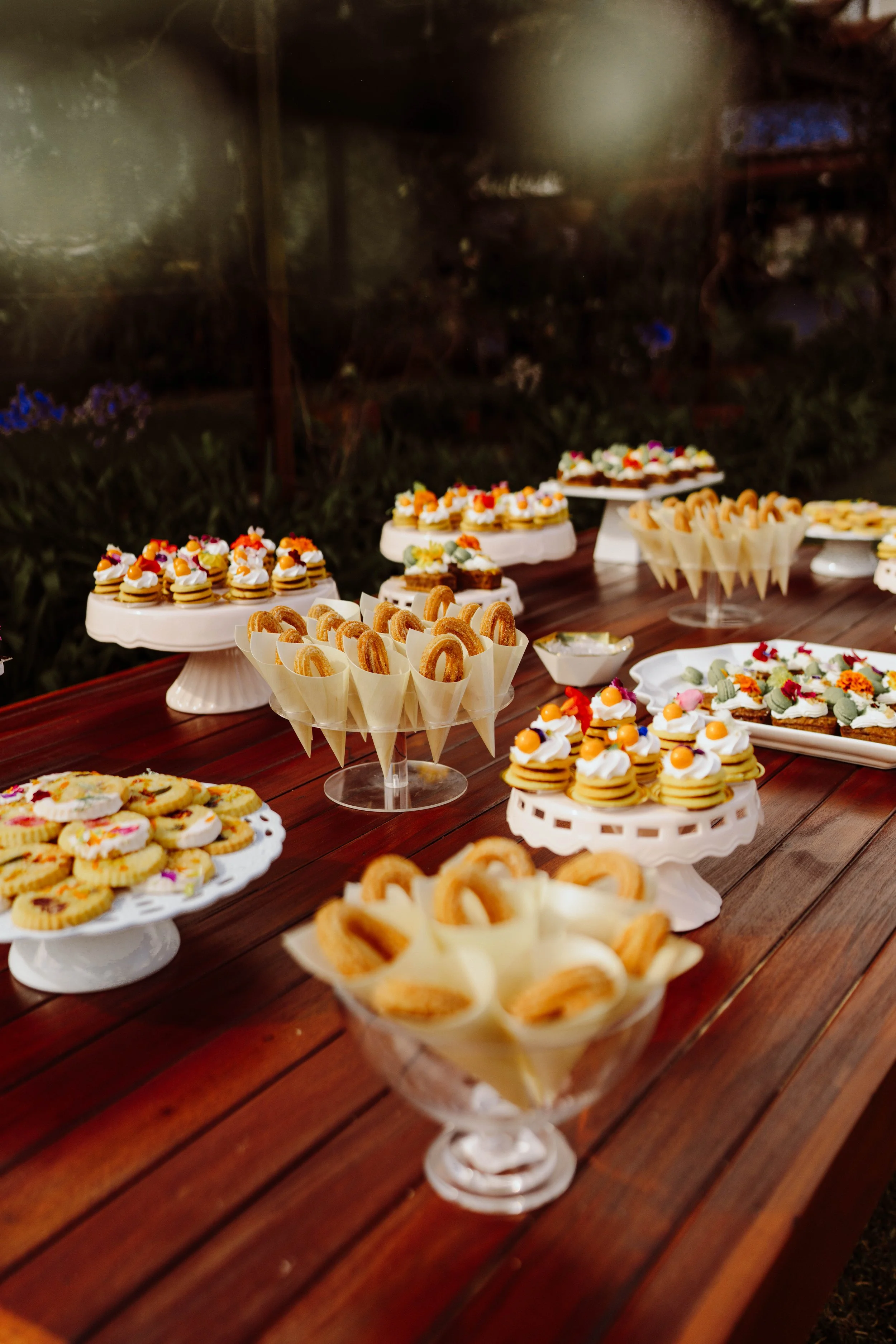 Mesa de postres decorada con galletas, mini pasteles y dulces sobre bases blancas y de vidrio durante una boda al aire libre.