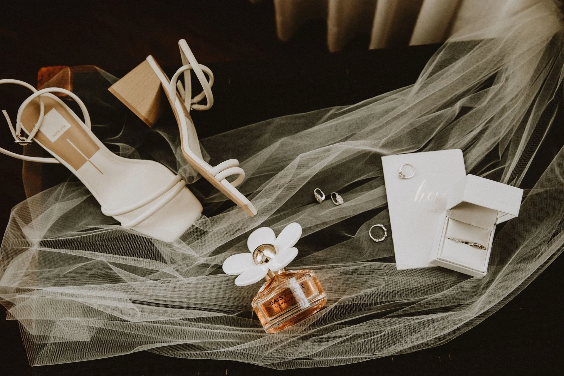 Wedding accessories including a pair of white high-heeled shoes, rings, earrings, perfume, and a white flower, arranged on black fabric with overlaying tulle fabric.
