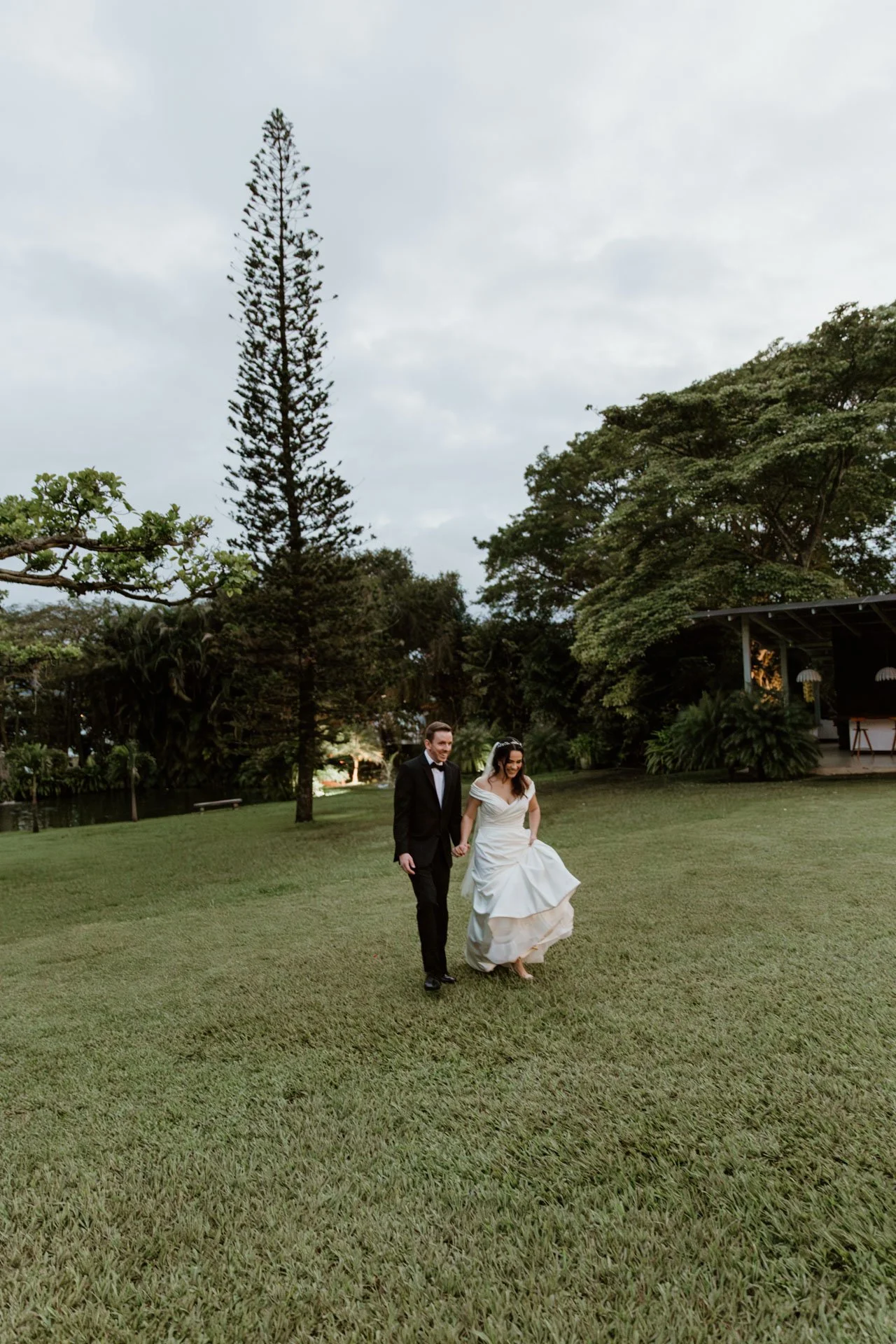 A bride and groom walking hand in hand across a grassy lawn outdoors during daytime with trees and a pavilion in the background.