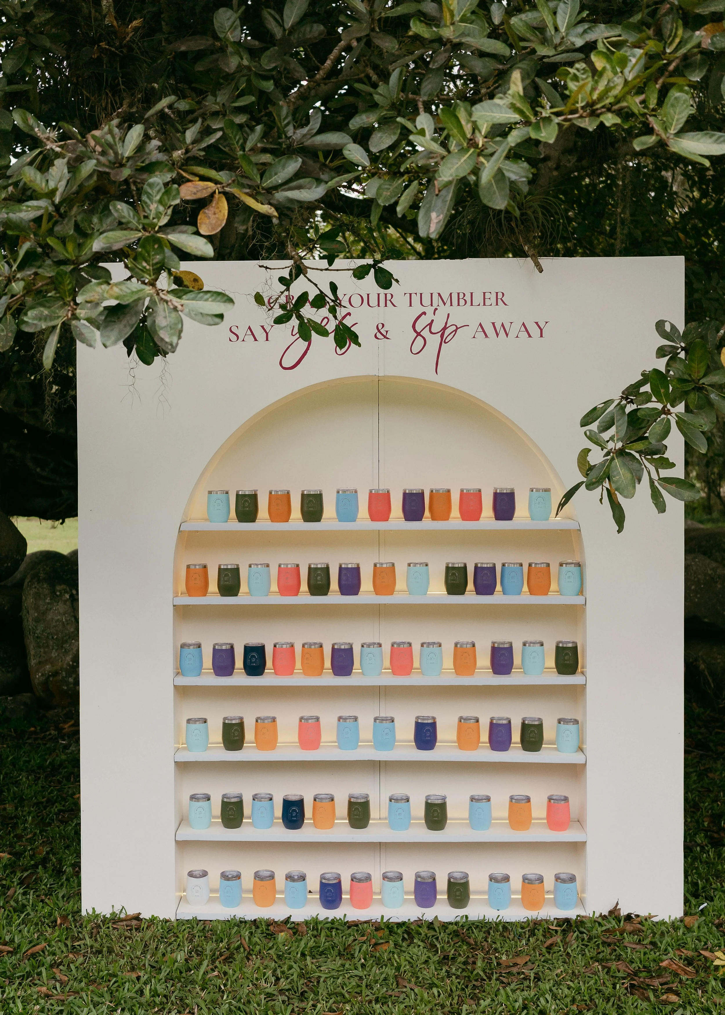 Colorful tumblers on a white display shelf outdoors with trees and grass in the background.