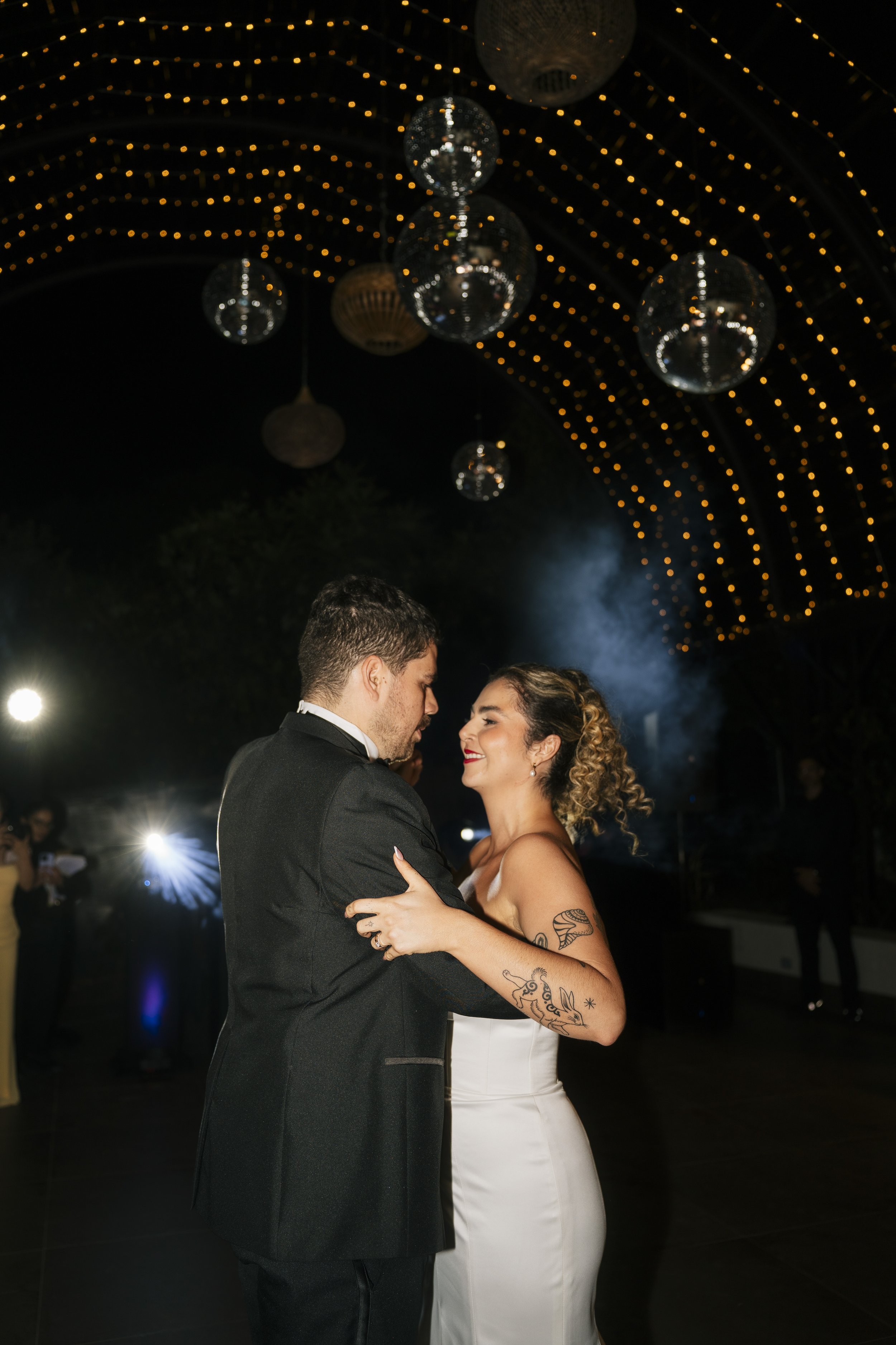 Novios bailando durante la fiesta de su boda, con iluminación cálida y detalles decorativos suspendidos sobre la pista.