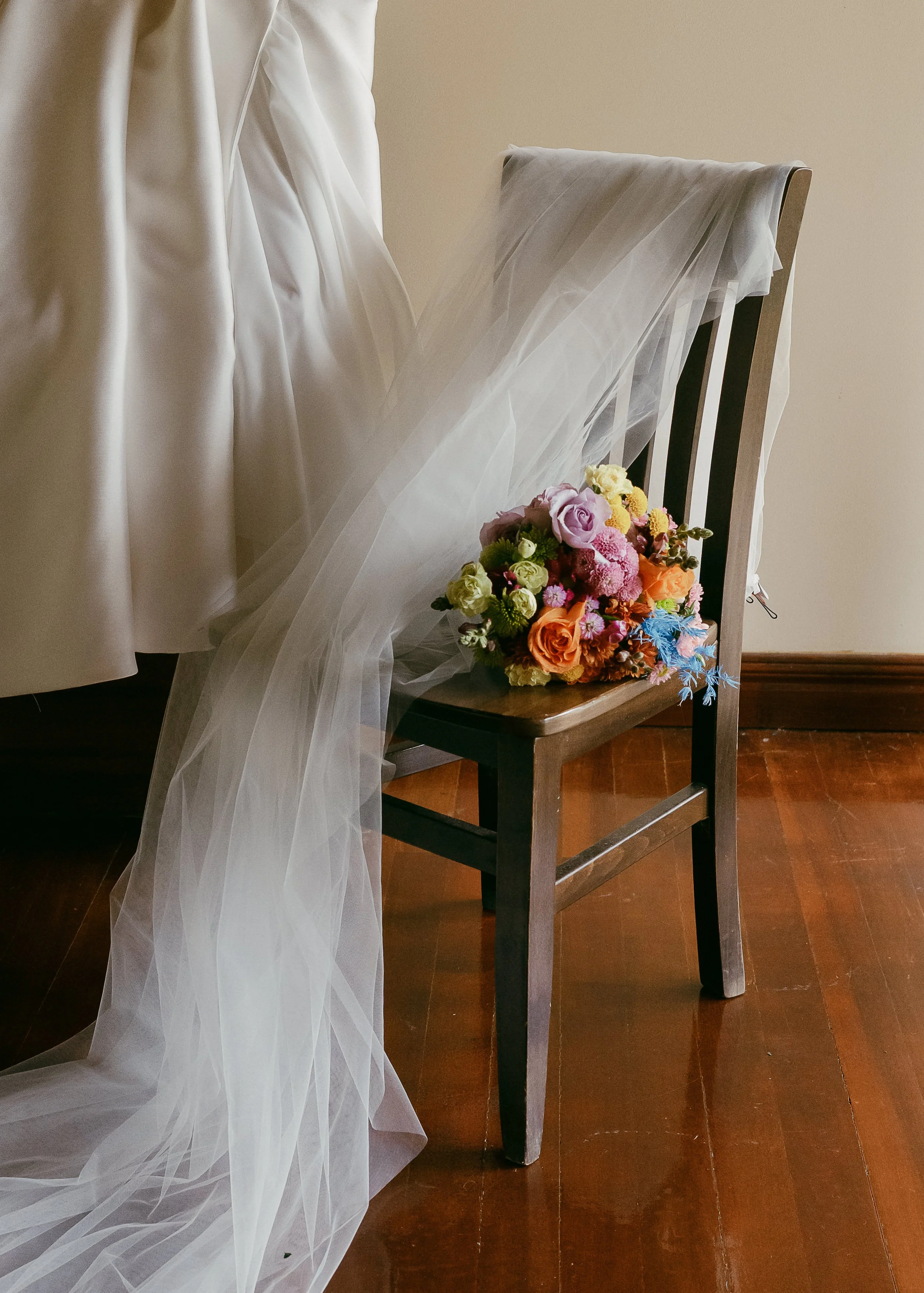 A bouquet of colorful flowers resting on a wooden chair beside a white wedding dress and veil.