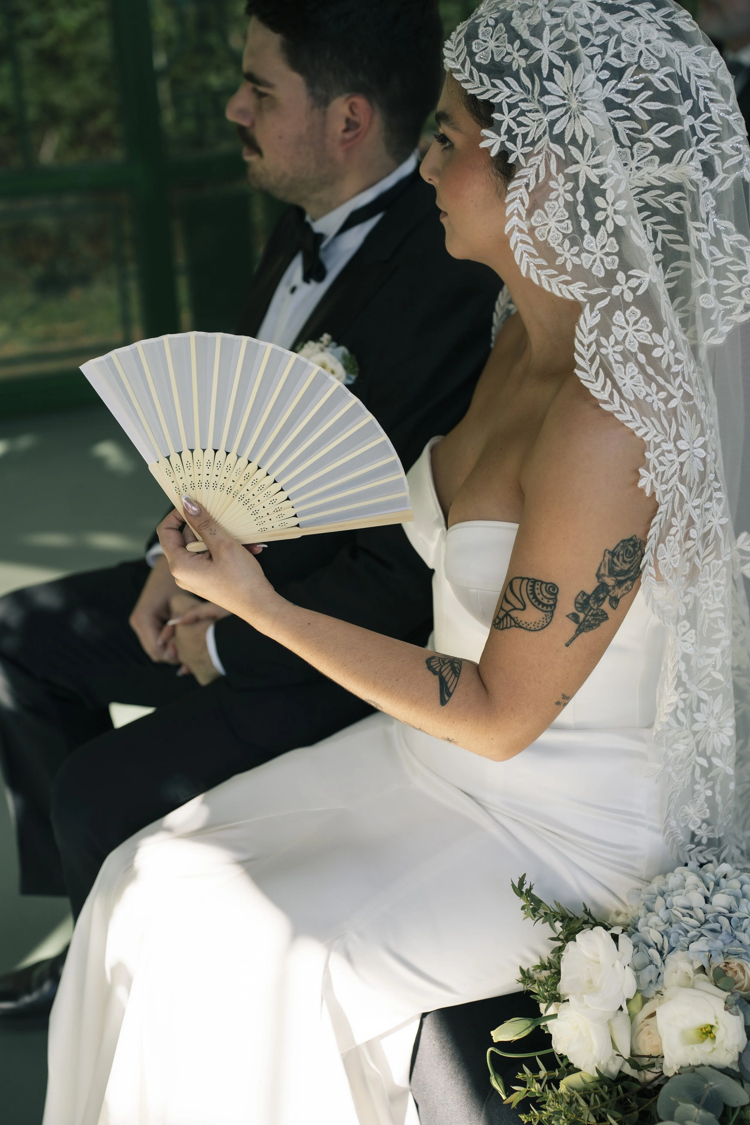 Pareja sentada durante la ceremonia de su boda, con la novia sosteniendo un abanico y detalles florales en primer plano.