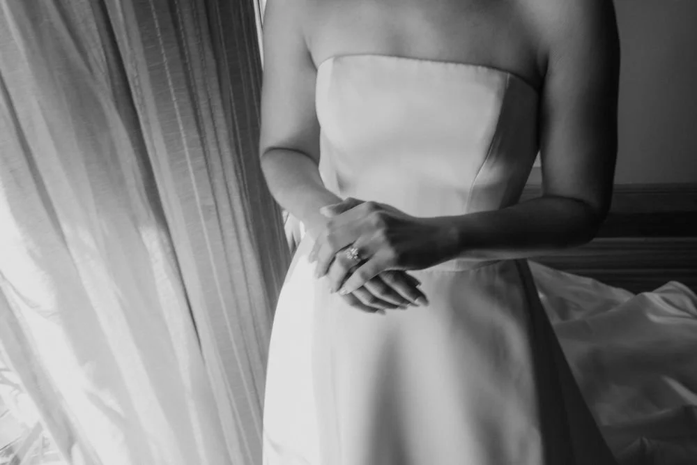 A woman in a strapless dress standing by a window with curtains, with her hands clasped in front of her.