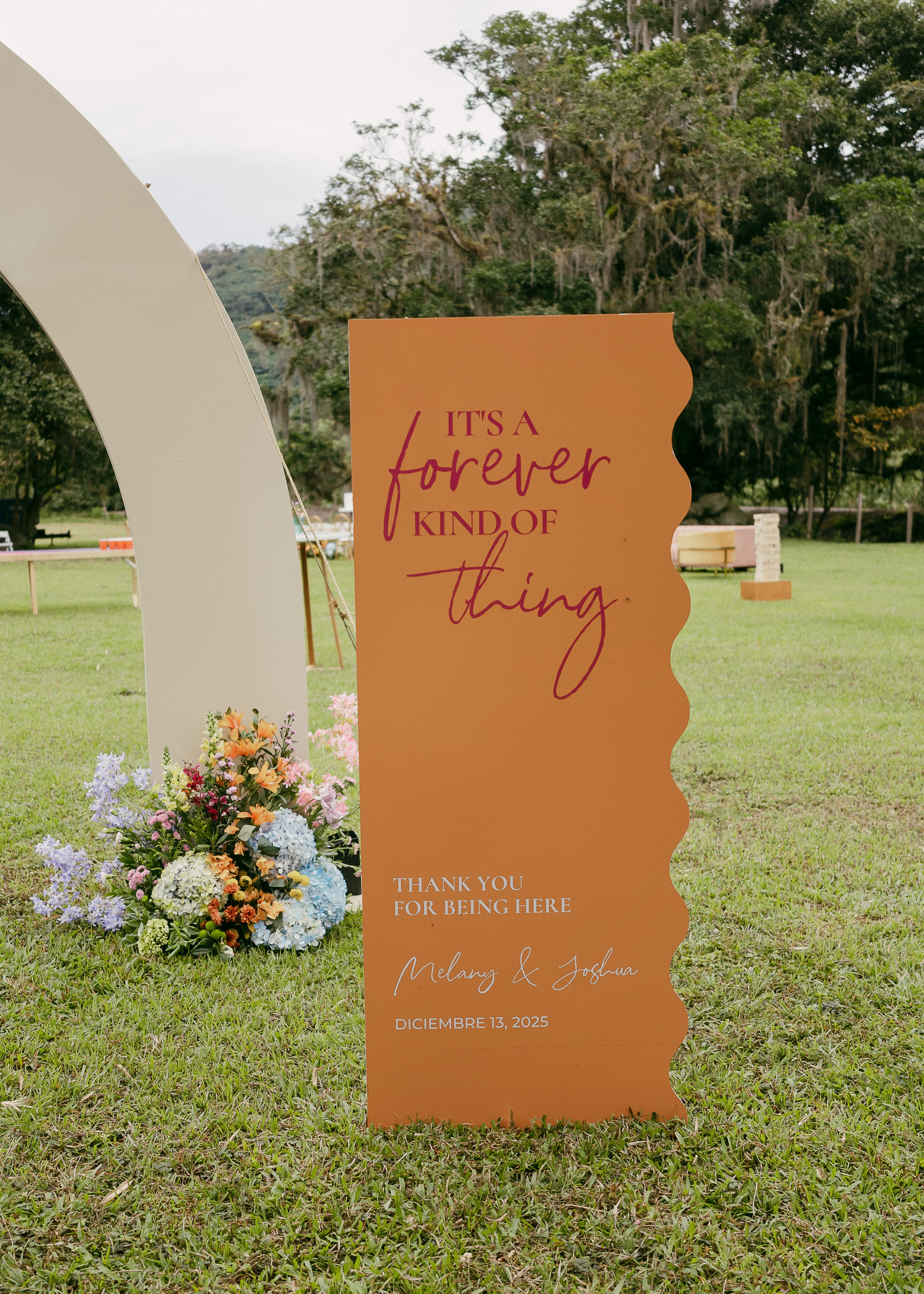 A wedding sign on an outdoor grassy area that reads, 'It's a forever kind of thing. Thank you for being here. Melany & Joshua, December 13, 2025.' There is a bouquet of flowers and a decorative arch in the background.