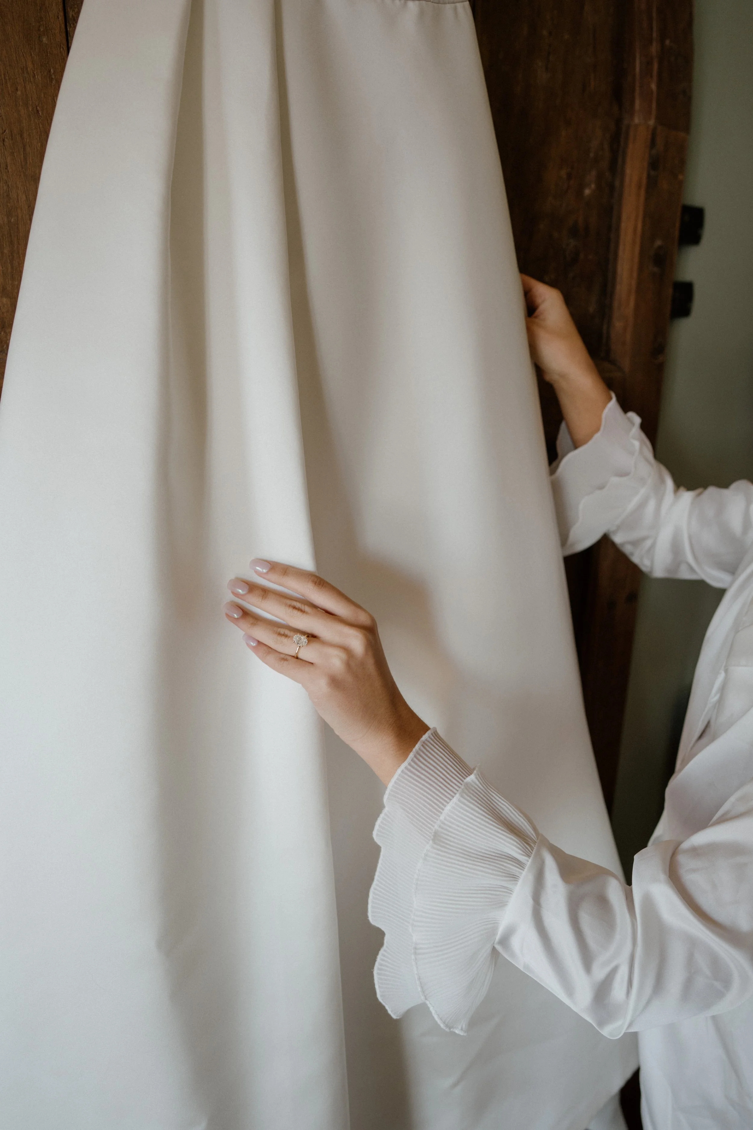A bride to be in a white shirt with pleated cuffs is touching a cream-colored fabric, possibly a wedding dress or gown, hanging on a wooden wall.