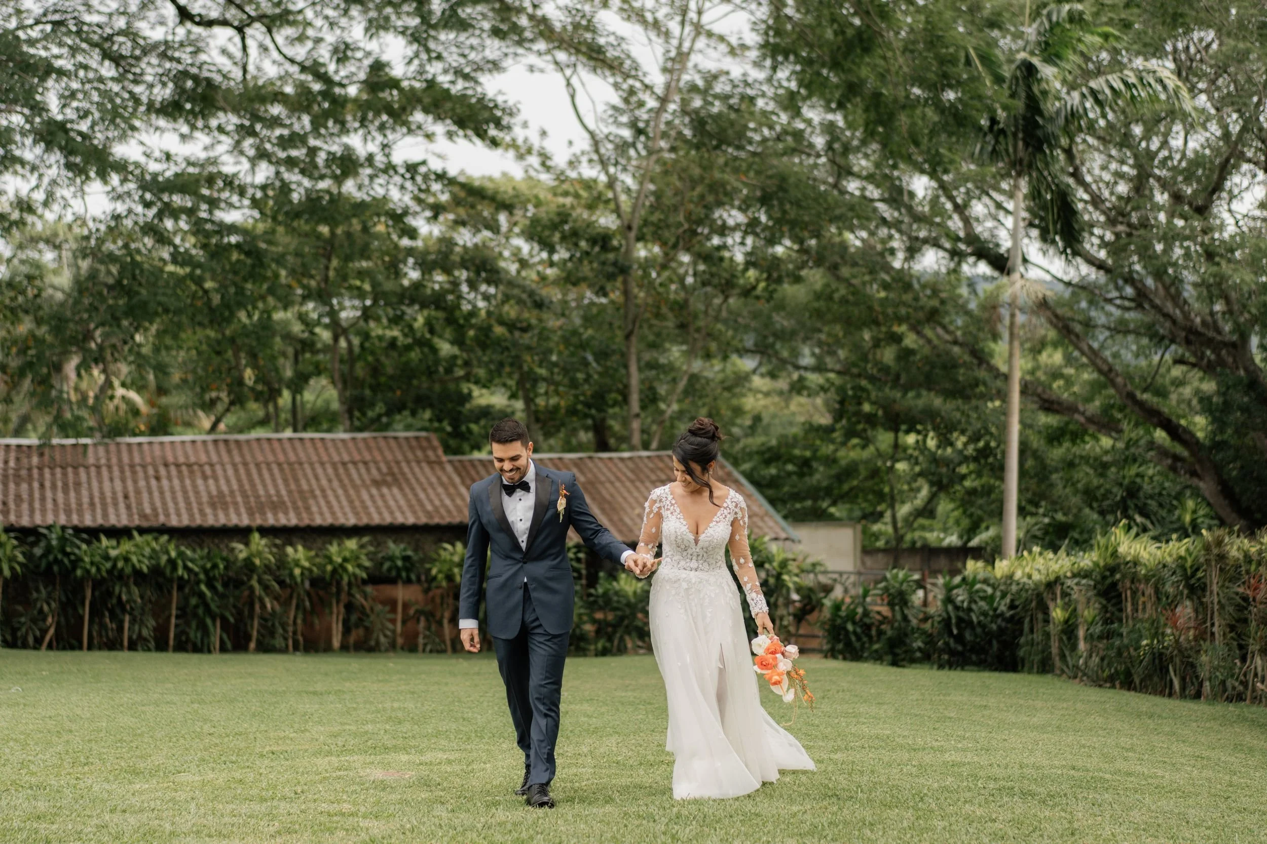 A newlywed couple walking on a green lawn, holding hands. The groom is dressed in a navy blue tuxedo with a black bowtie. The bride is wearing a white wedding gown with lace details and is holding a bouquet of pink and orange flowers. They are outdoors, surrounded by trees and a rustic building in the background.