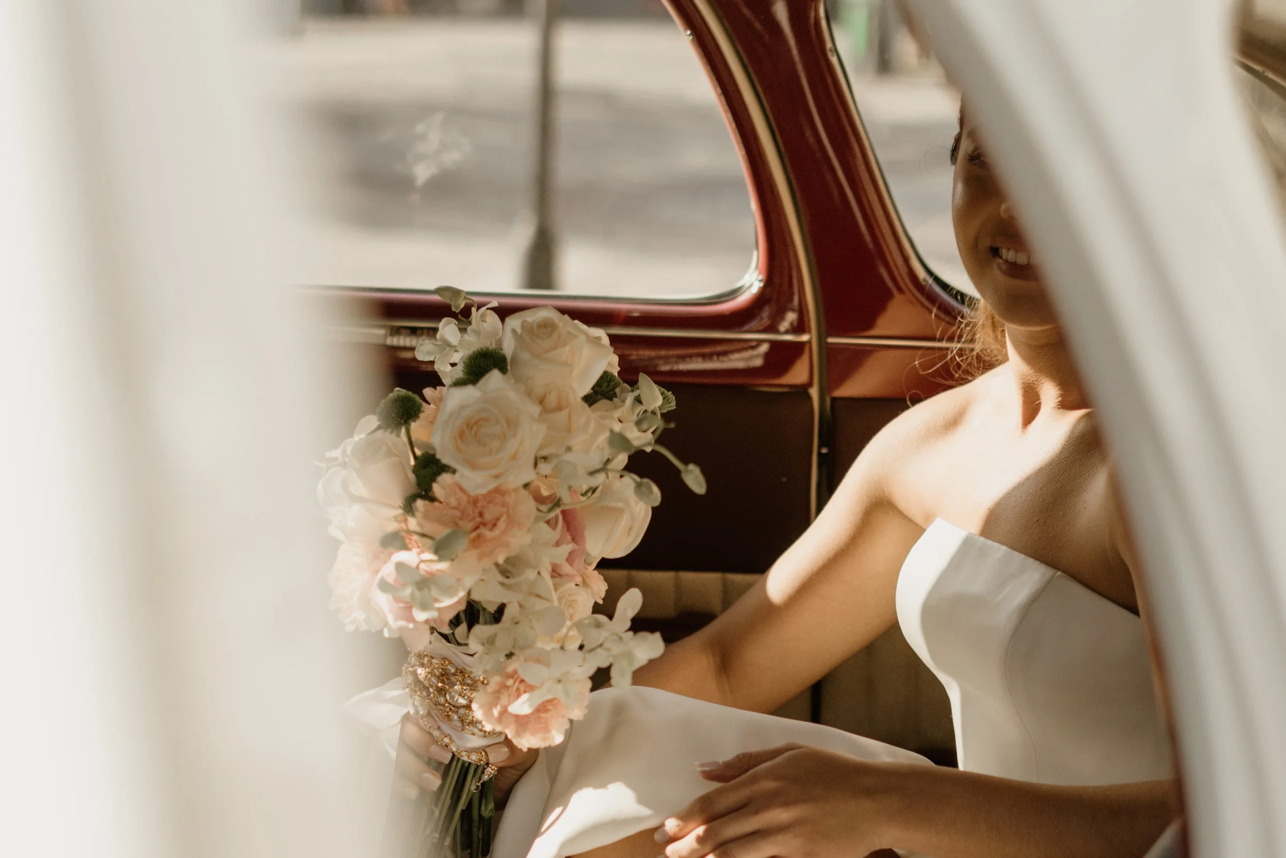 A young woman in a white strapless dress sitting inside a vintage car, holding a bouquet of white and pink flowers, with her face partially obscured by the car window frame.