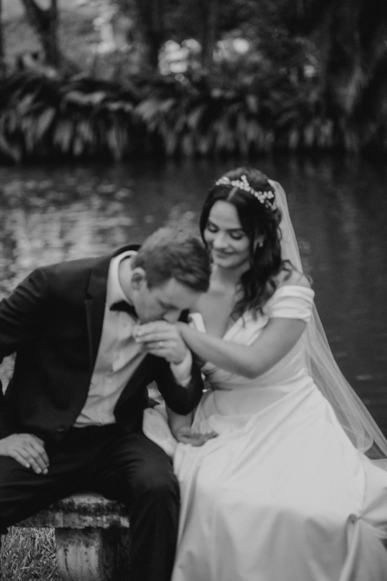 A black and white photo of a bride and groom near a body of water. The groom is kissing the bride's hand, who is seated and smiling, wearing a wedding dress and veil.