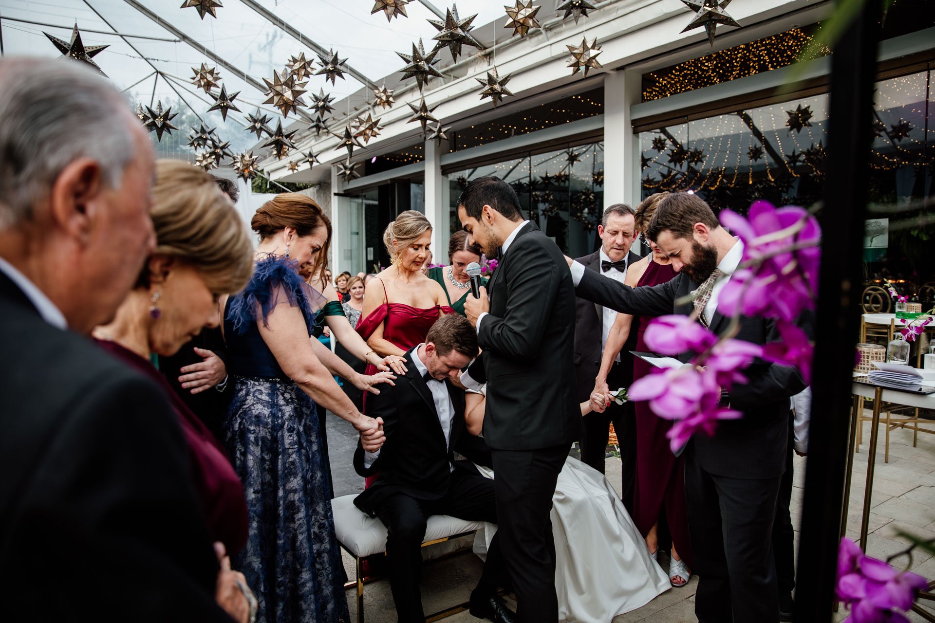 Momento de oración durante la ceremonia de boda, con los novios arrodillados y acompañados por personas cercanas en un espacio decorado con luces y estrellas suspendidas.
